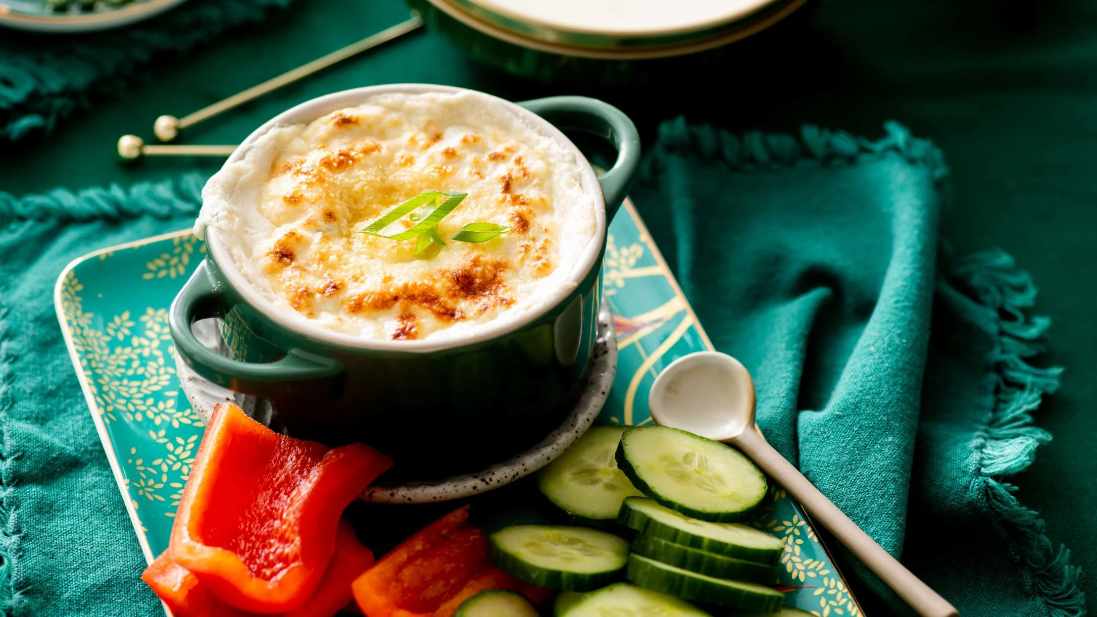 Creamy baked dip in teal ramekin garnished with green onions, served with cucumber slices and red pepper on turquoise cloth.