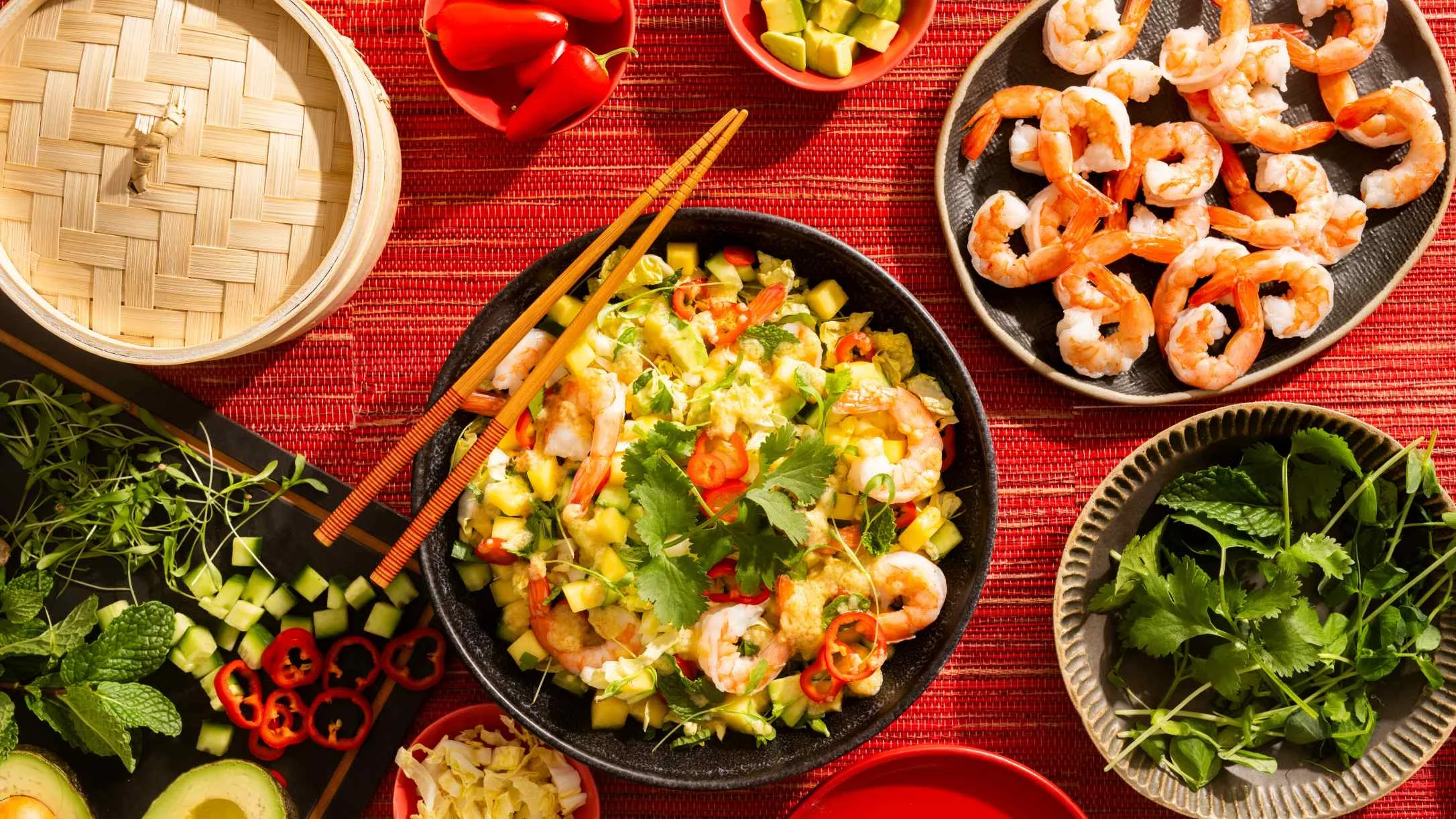 Asian-style shrimp dish with colorful vegetables on red placemat, surrounded by fresh herbs, avocado, and cooked shrimp.