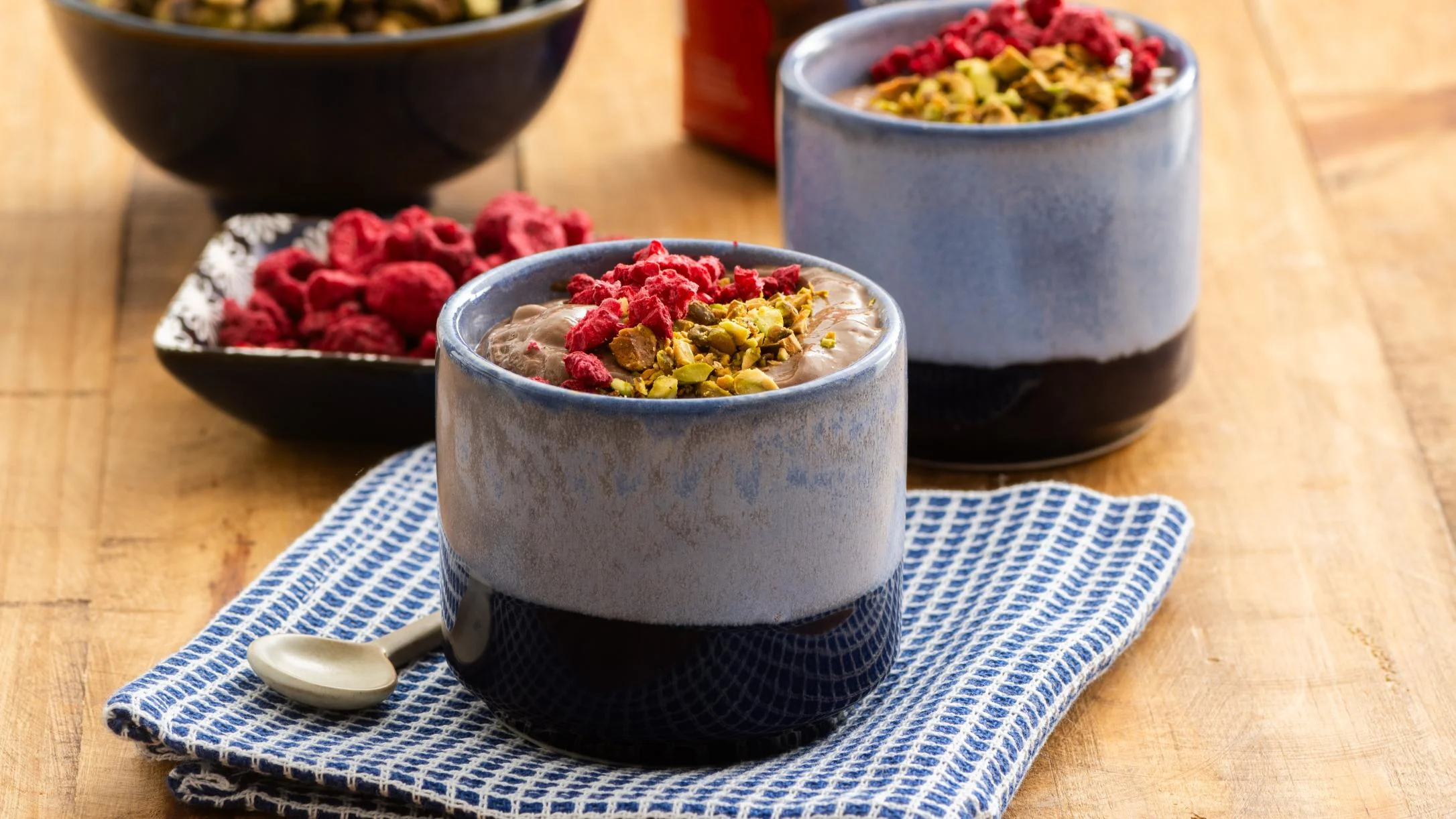 Blue ceramic cups filled with pudding topped with pistachios and dried raspberries on a striped cloth napkin.