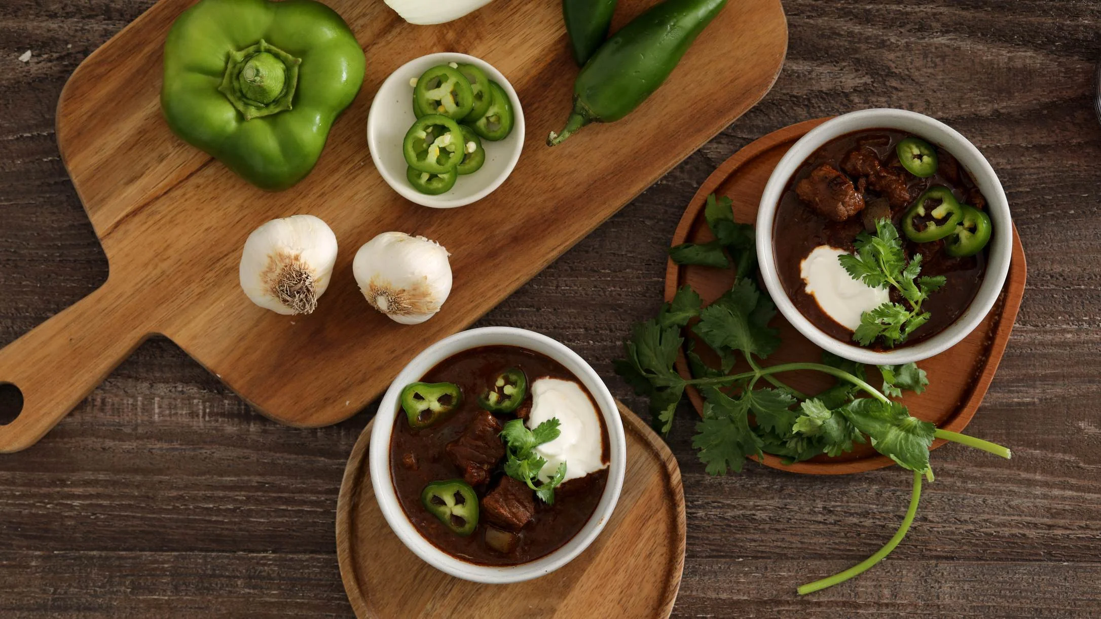 Two bowls of beef chili topped with sour cream, jalapeños, and cilantro, alongside fresh peppers and garlic on a wooden board.