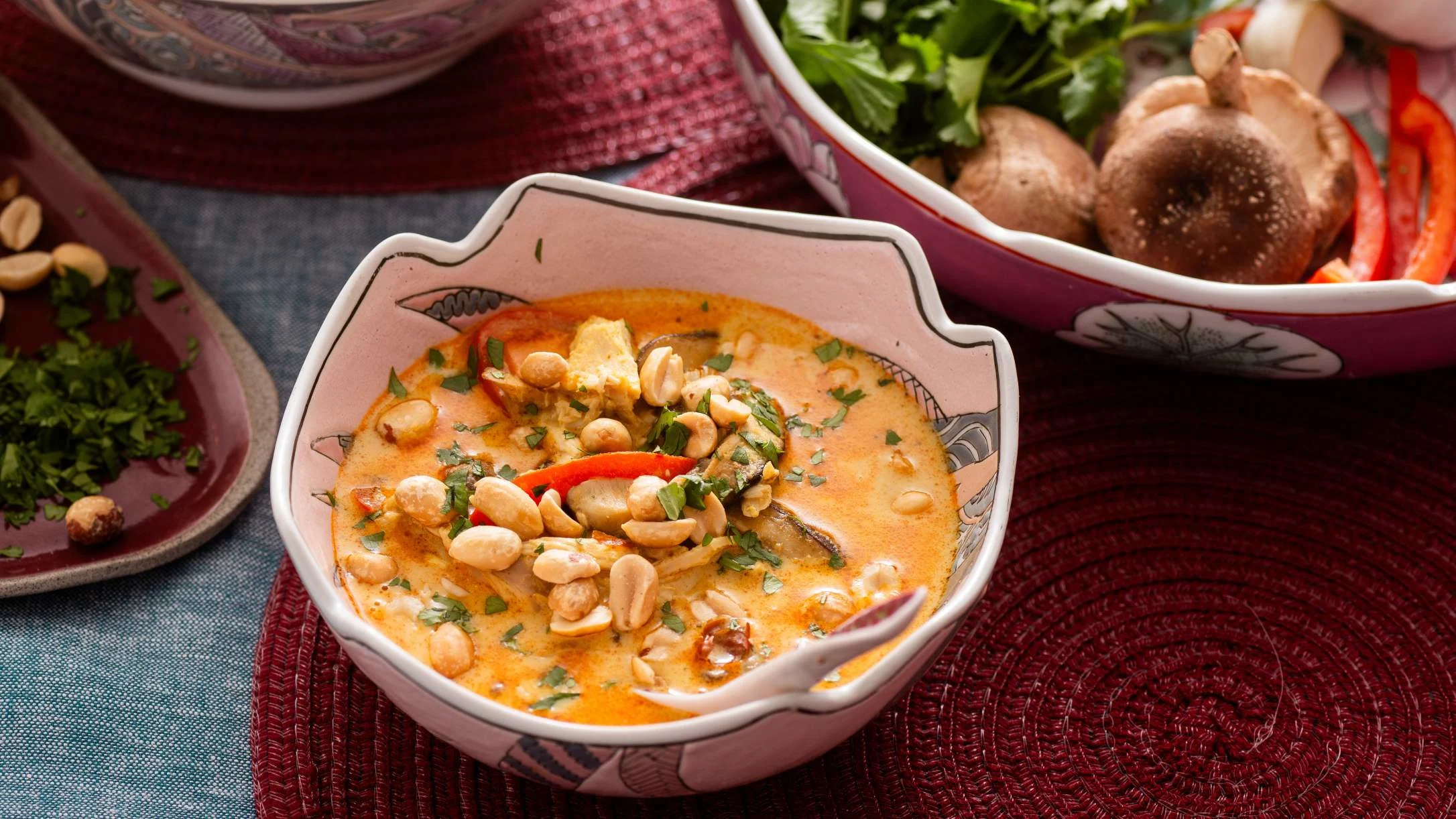 Creamy orange curry with peanuts and herbs in a decorative bowl, with fresh vegetables in background dishes.