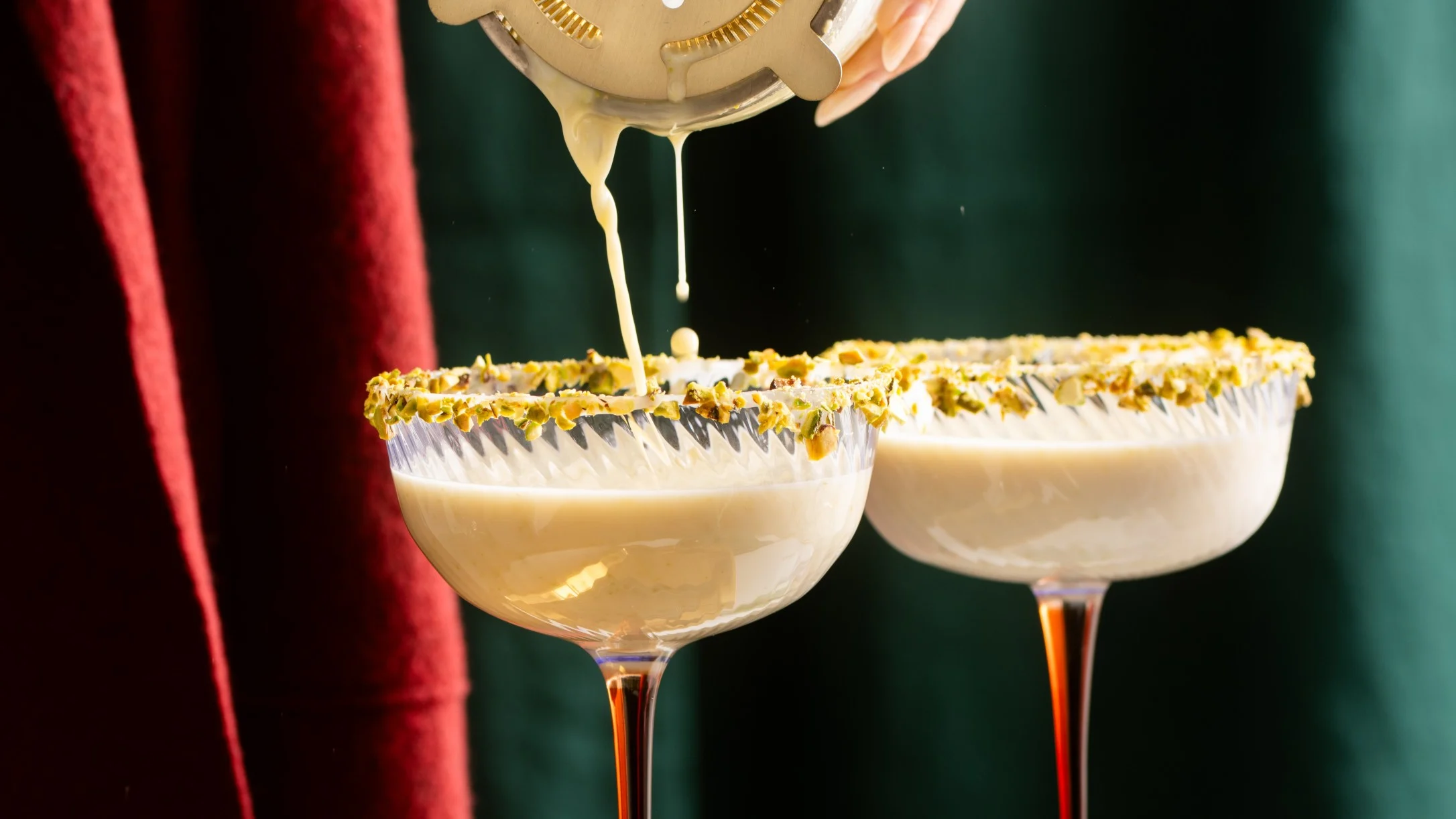 Two cocktail glasses with pistachio-crusted rims against a dark green background.