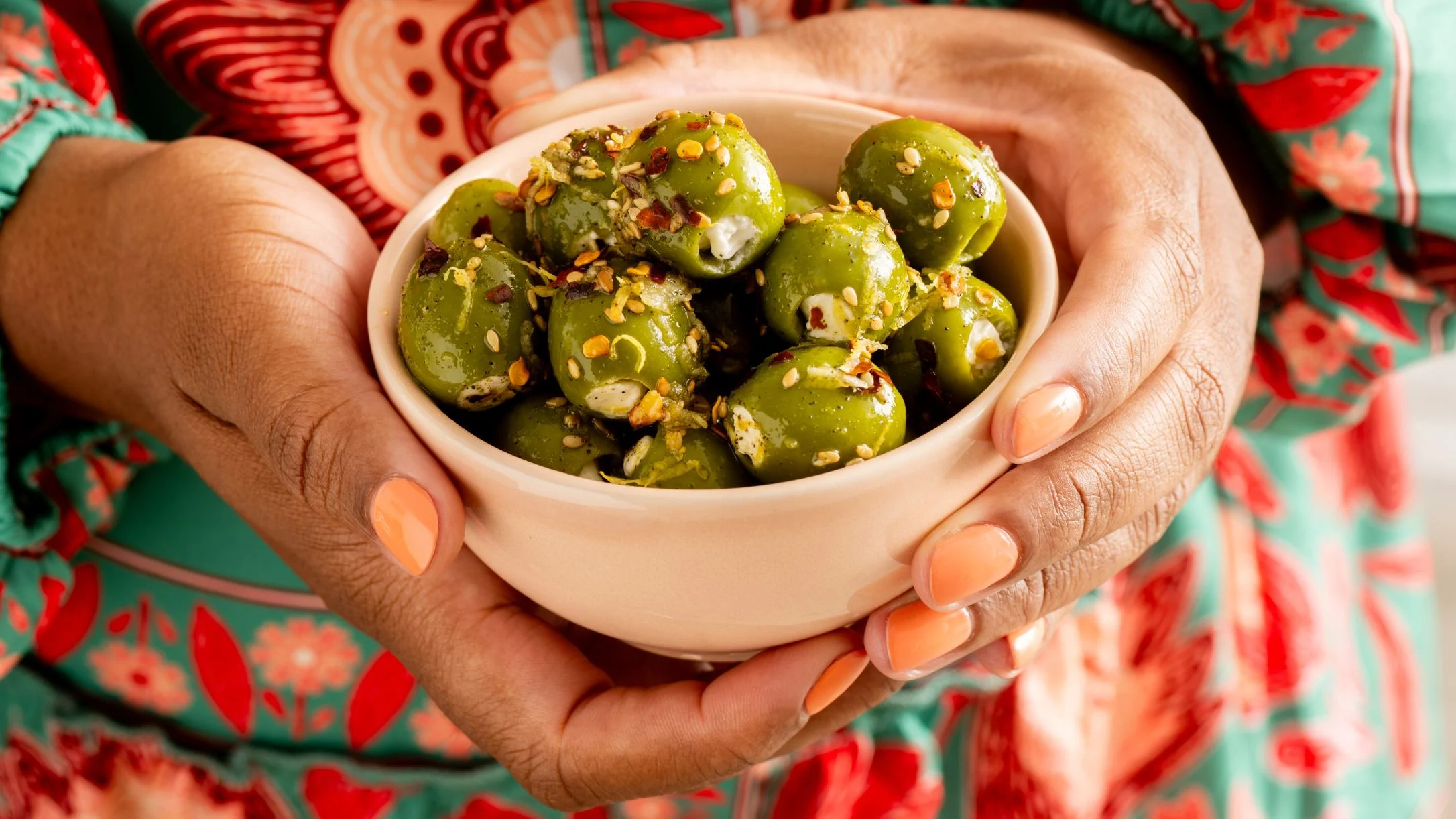 Hands with peach nails cradling a small bowl of seasoned green olives, wearing a colorful teal and red floral outfit.