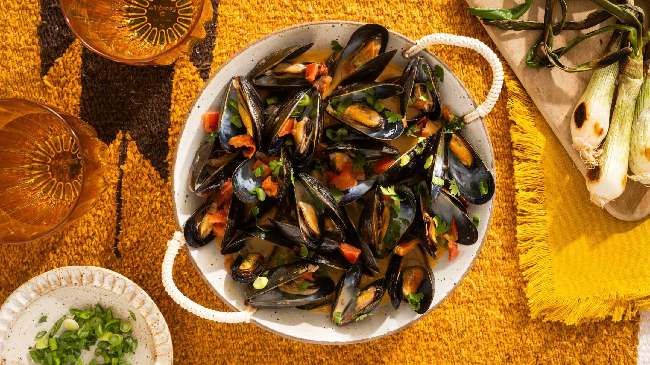 Grilled mussels in white ceramic pot with chipotle and herbs, served with wine glasses on yellow textured tablecloth.