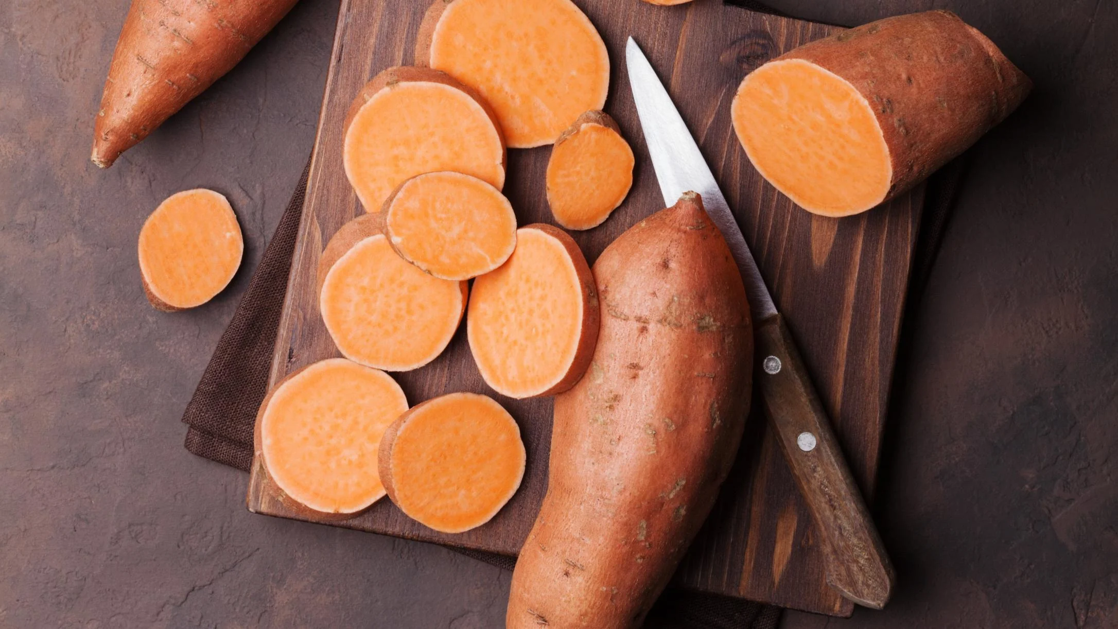 Sliced sweet potatoes with bright orange flesh on a wooden cutting board, alongside whole sweet potatoes and a knife.