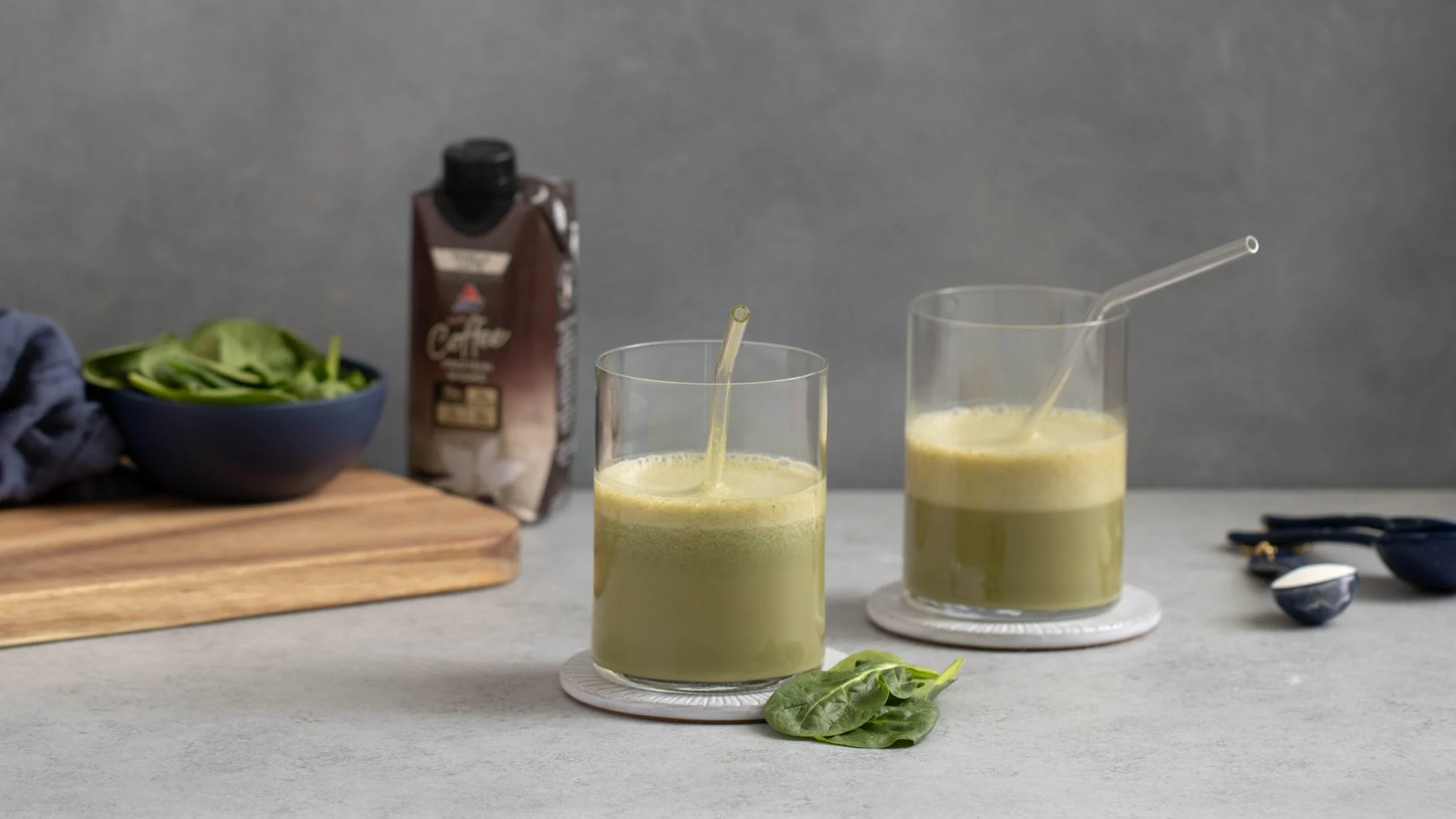 Two glasses of green smoothie with straws, spinach leaves, a cutting board, and an Atkins coffee drink on a gray surface.