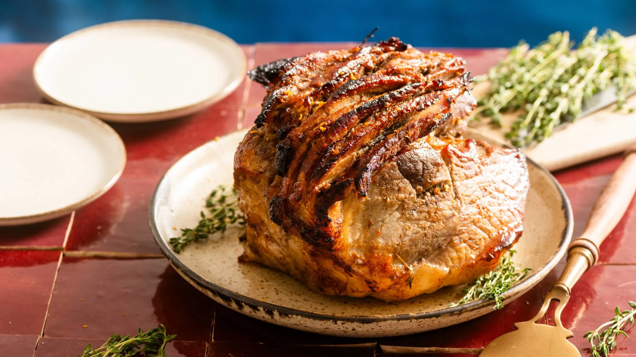 Glazed roast ham with crispy scored top on ceramic plate, garnished with fresh herbs on red tile table.