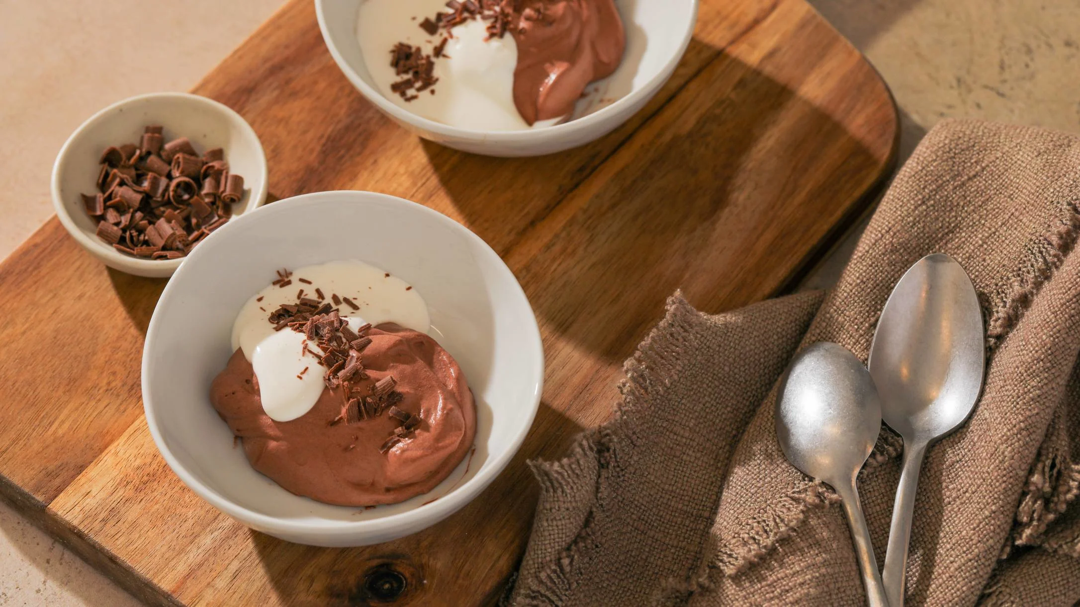 Two white bowls of chocolate mousse topped with cream and chocolate shavings on a wooden board with spoons.