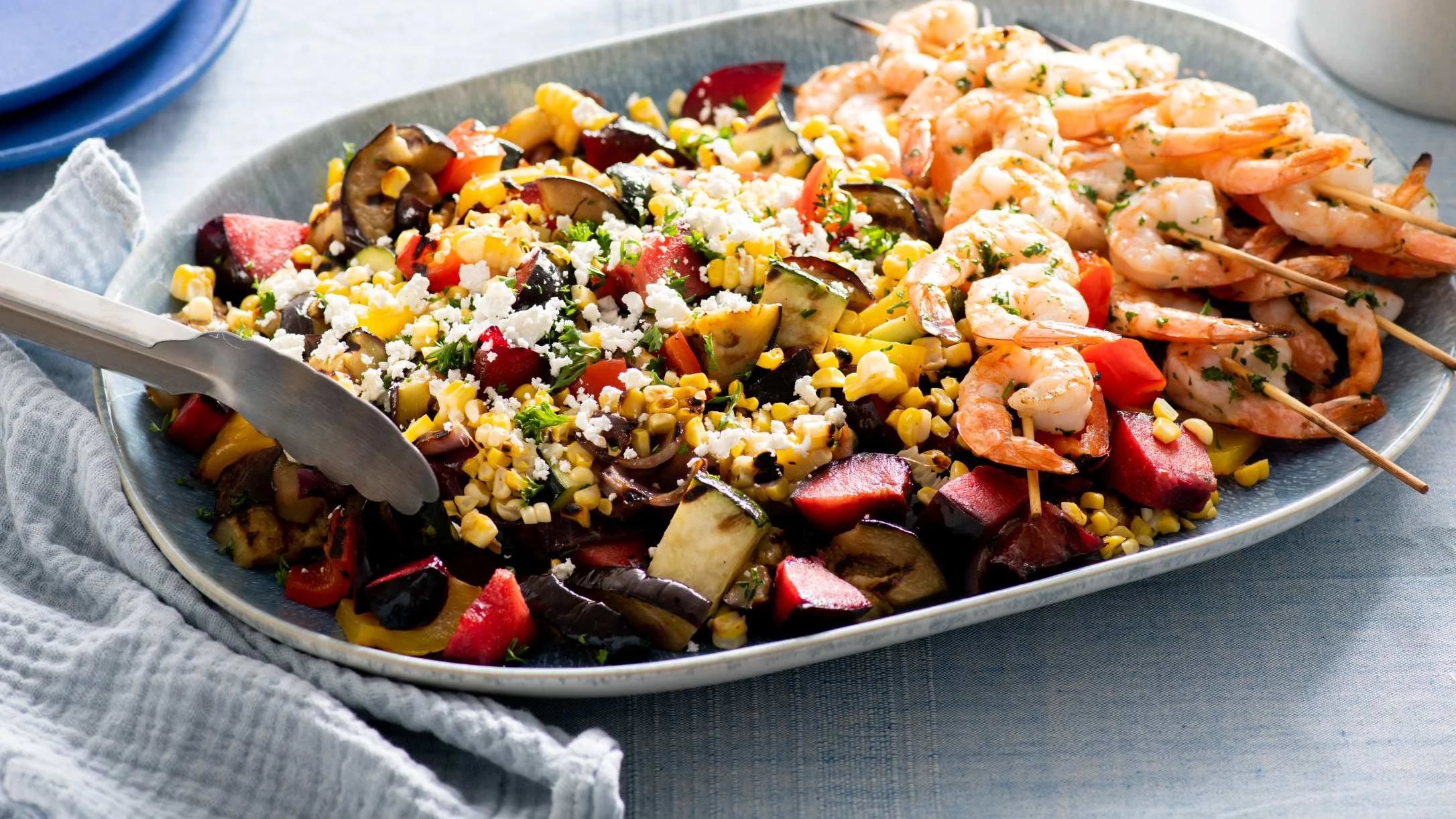 A platter of grilled shrimp skewers with colorful roasted vegetables, corn, crumbled cheese, and fresh herbs.
