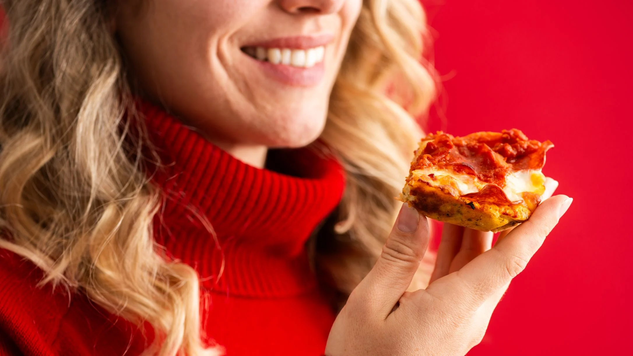 Person with blonde wavy hair in red sweater smiling while holding a slice of pizza against a bright red background.