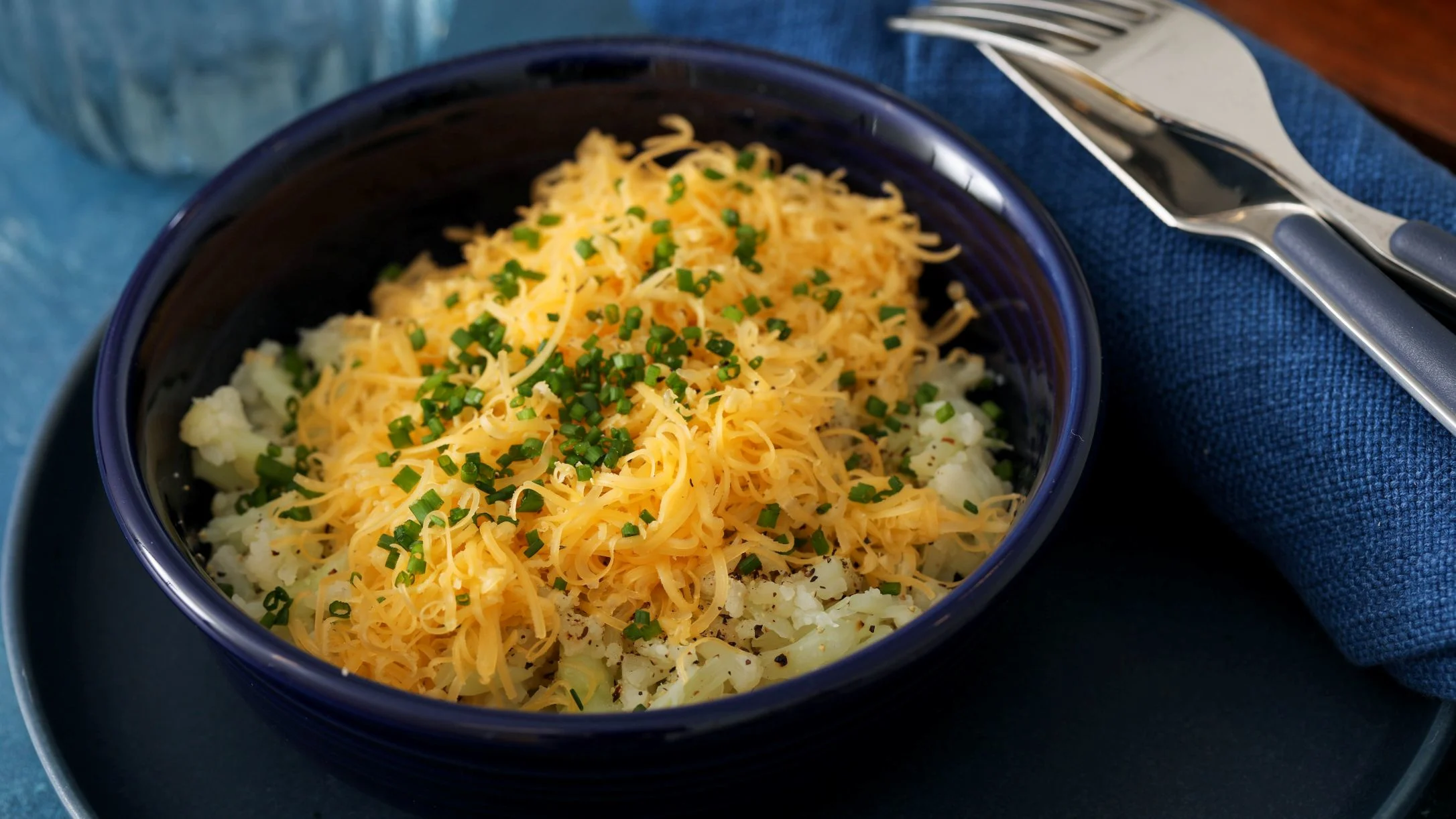Blue bowl containing cauliflower mash topped with shredded yellow cheese and chopped green chives, with silverware nearby.