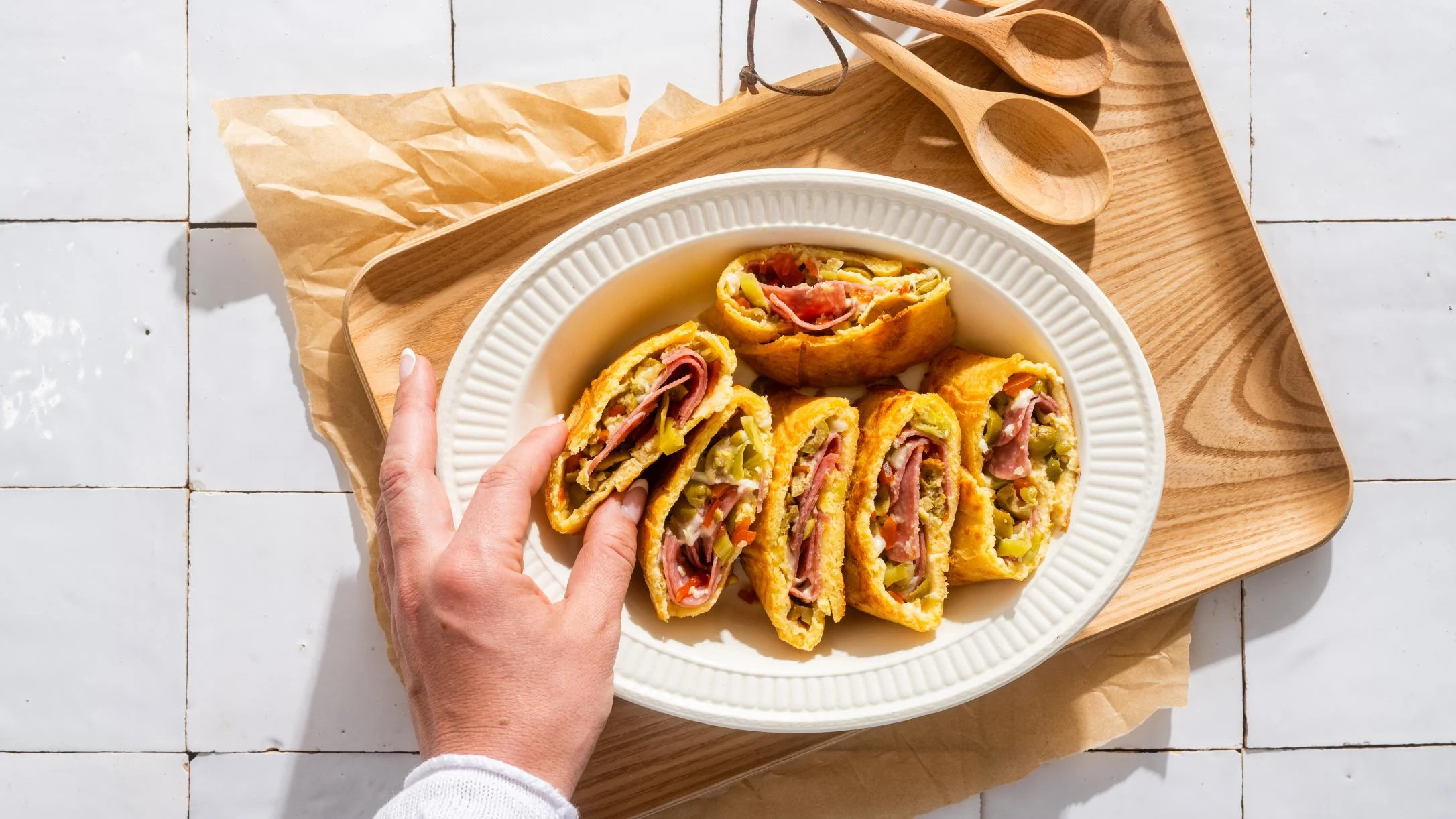 Hand reaching for sliced stromboli filled with ham and vegetables on white plate atop wooden serving board.