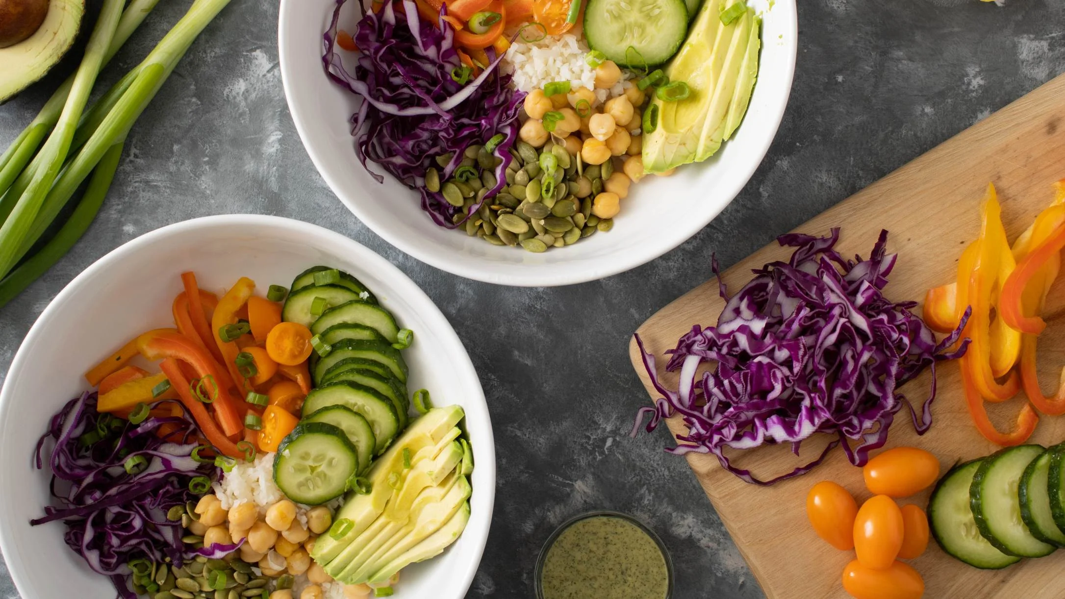 Two colorful veggie bowls with avocado, cucumber, cabbage, chickpeas, and peppers beside a cutting board with fresh ingredients.