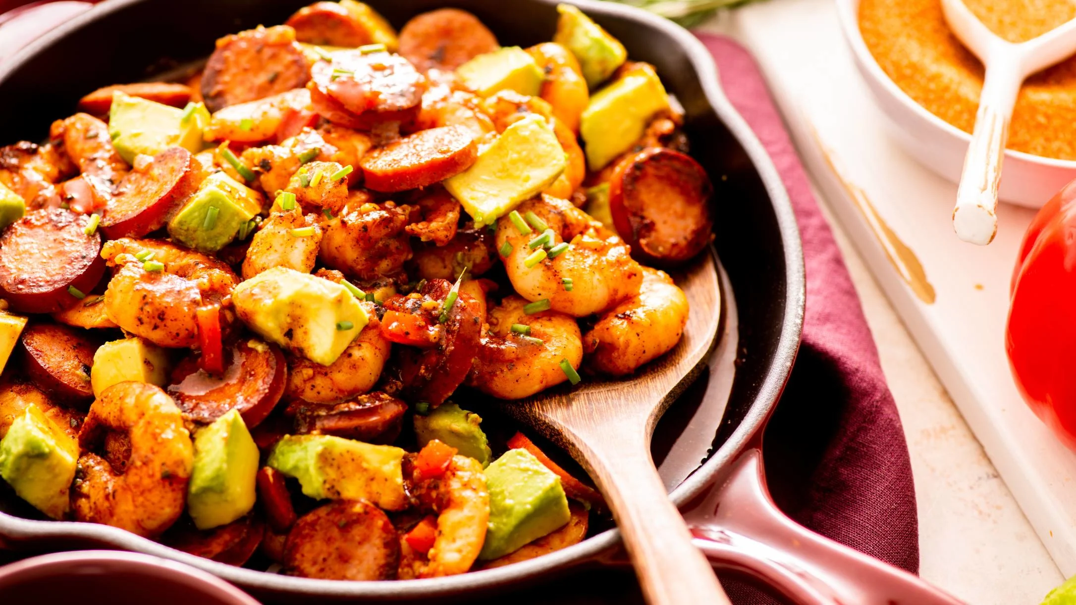 A cast iron skillet filled with seasoned shrimp, sliced sausage, and avocado chunks, garnished with chives.