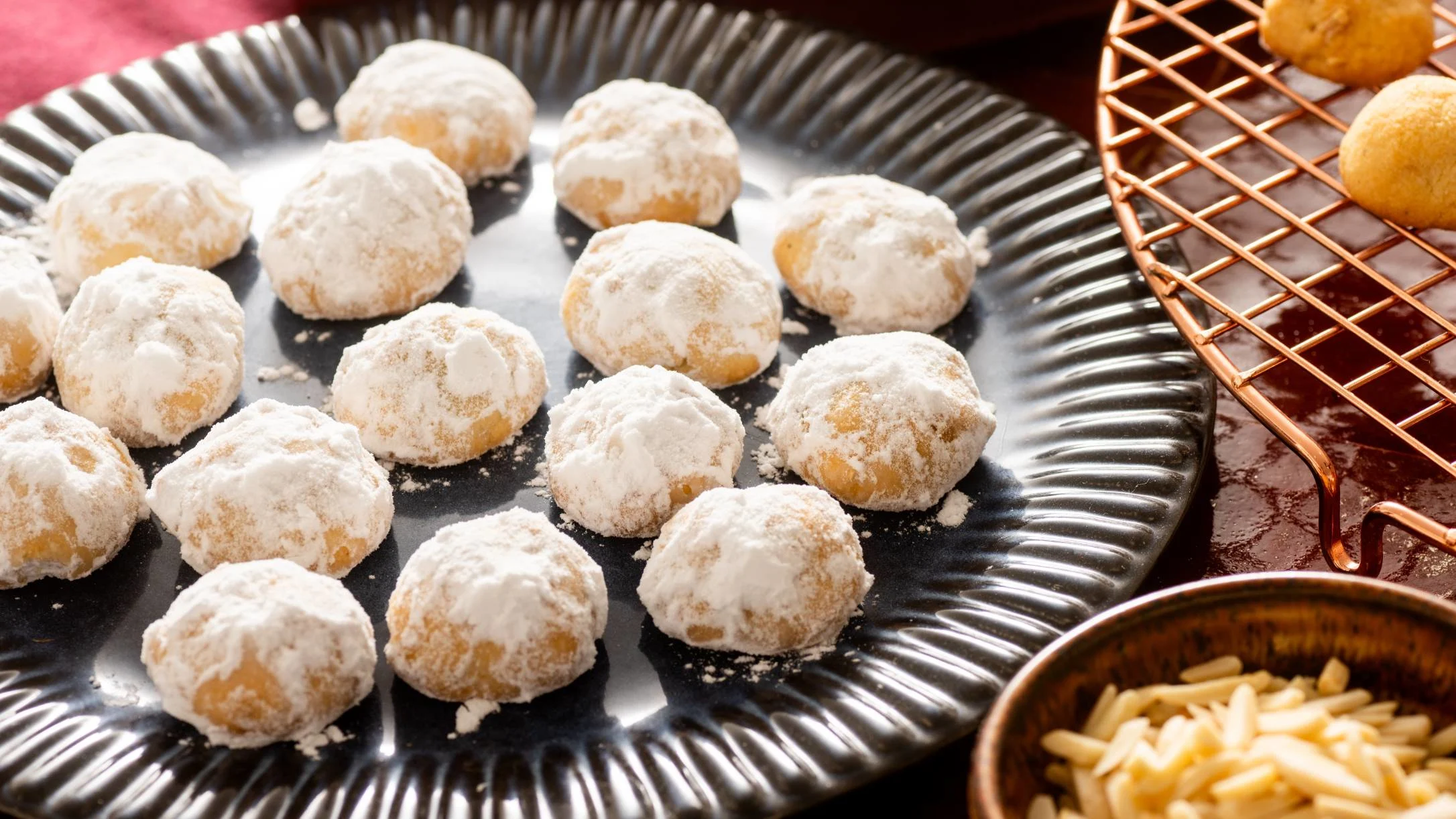 Powdered snowball cookies on a black plate with a copper cooling rack and small bowl of sliced almonds nearby.