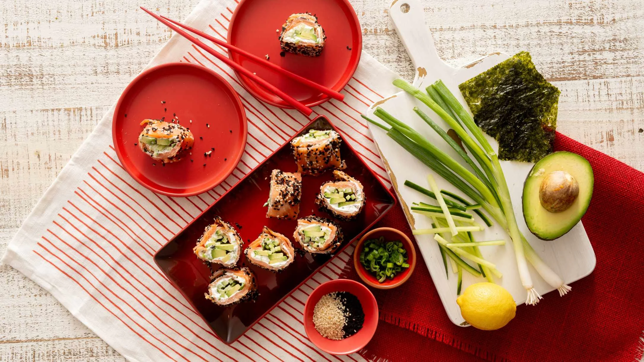 Sushi rolls with avocado and vegetables on red plates with chopsticks, alongside fresh ingredients on a white table.