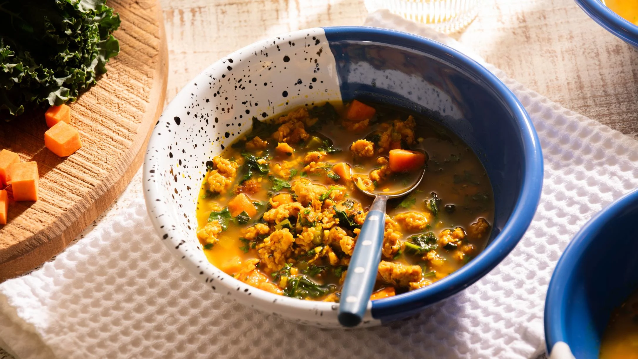 Hearty vegetable soup with ground meat and greens in a blue-rimmed speckled bowl with chopped sweet potato nearby.