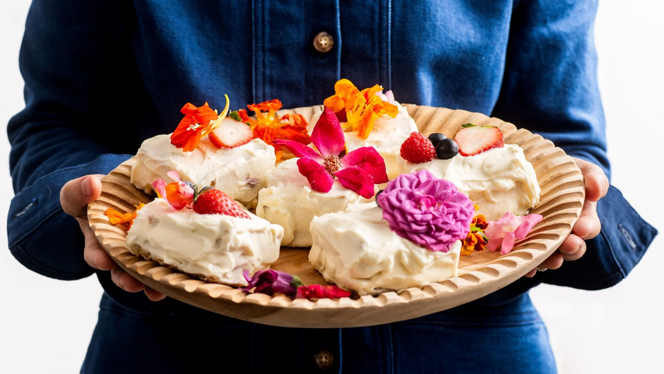 Person in blue denim shirt holding wooden plate with cream desserts decorated with colorful edible flowers and berries.