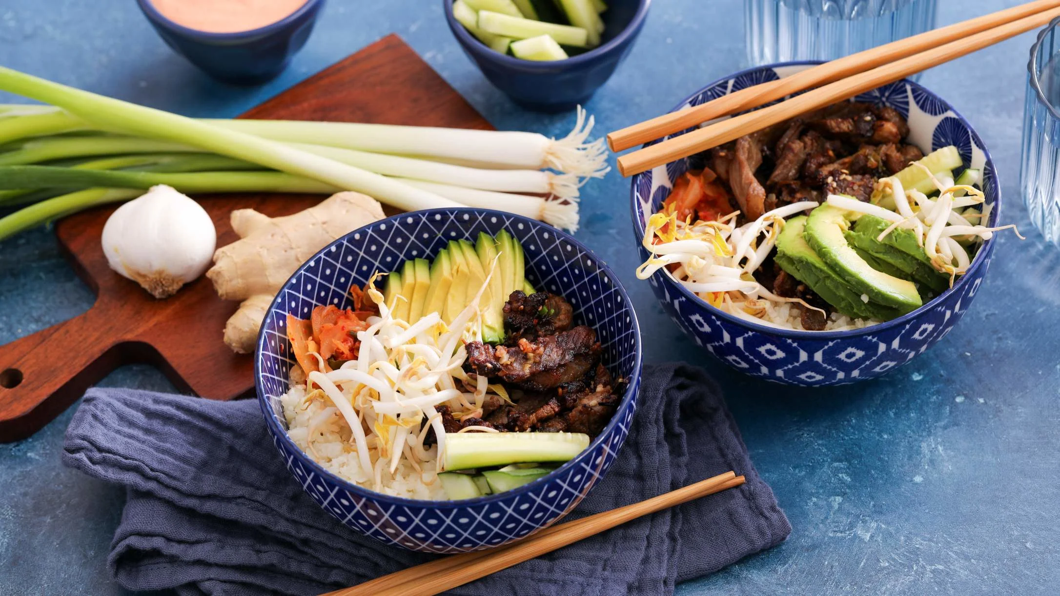 Two blue patterned bowls with rice, grilled meat, avocado, bean sprouts, and veggies, with chopsticks, scallions, and ginger nearby.