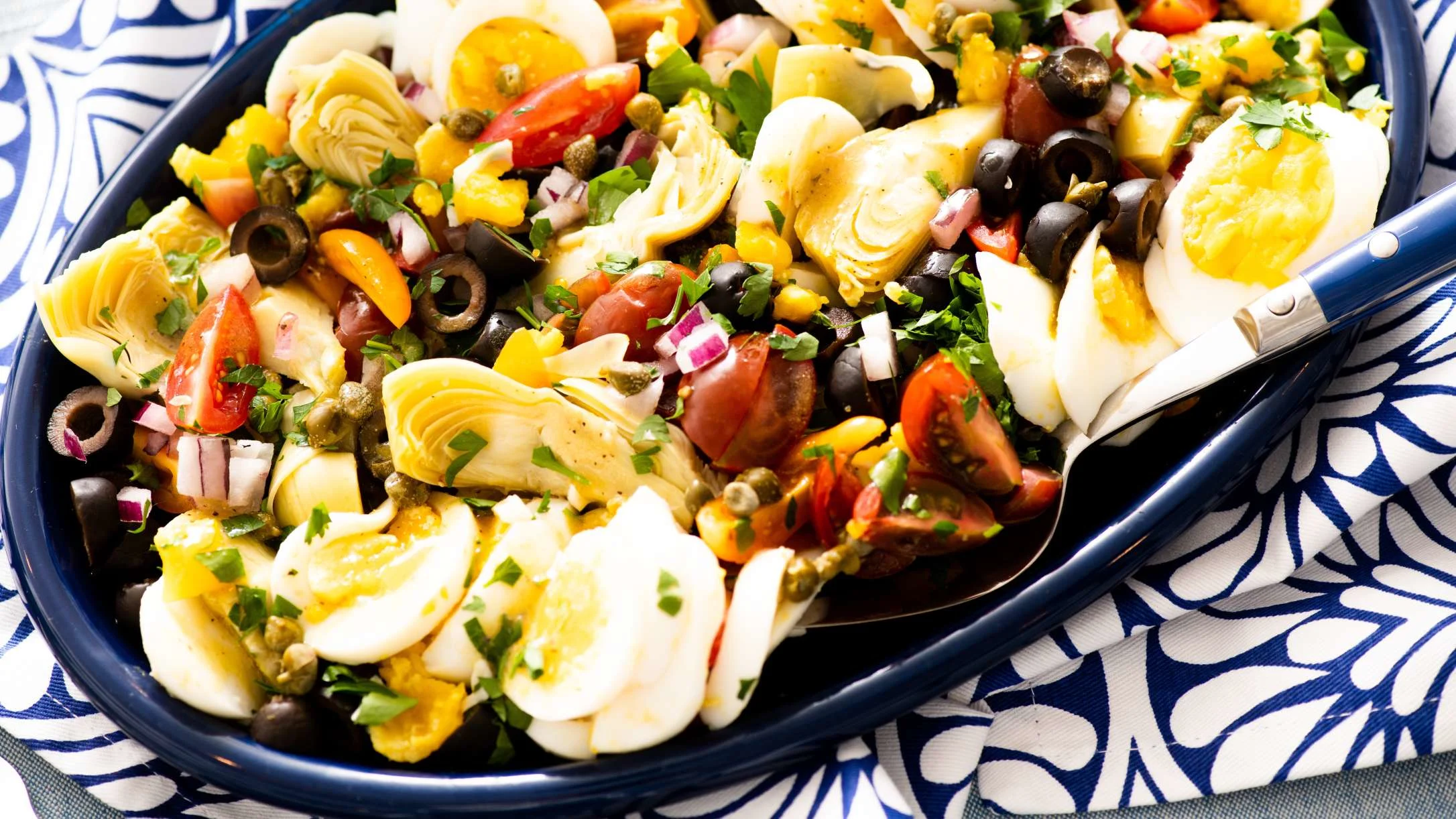 Mediterranean salad with hard-boiled eggs, artichokes, olives, tomatoes, and capers in a blue serving dish.