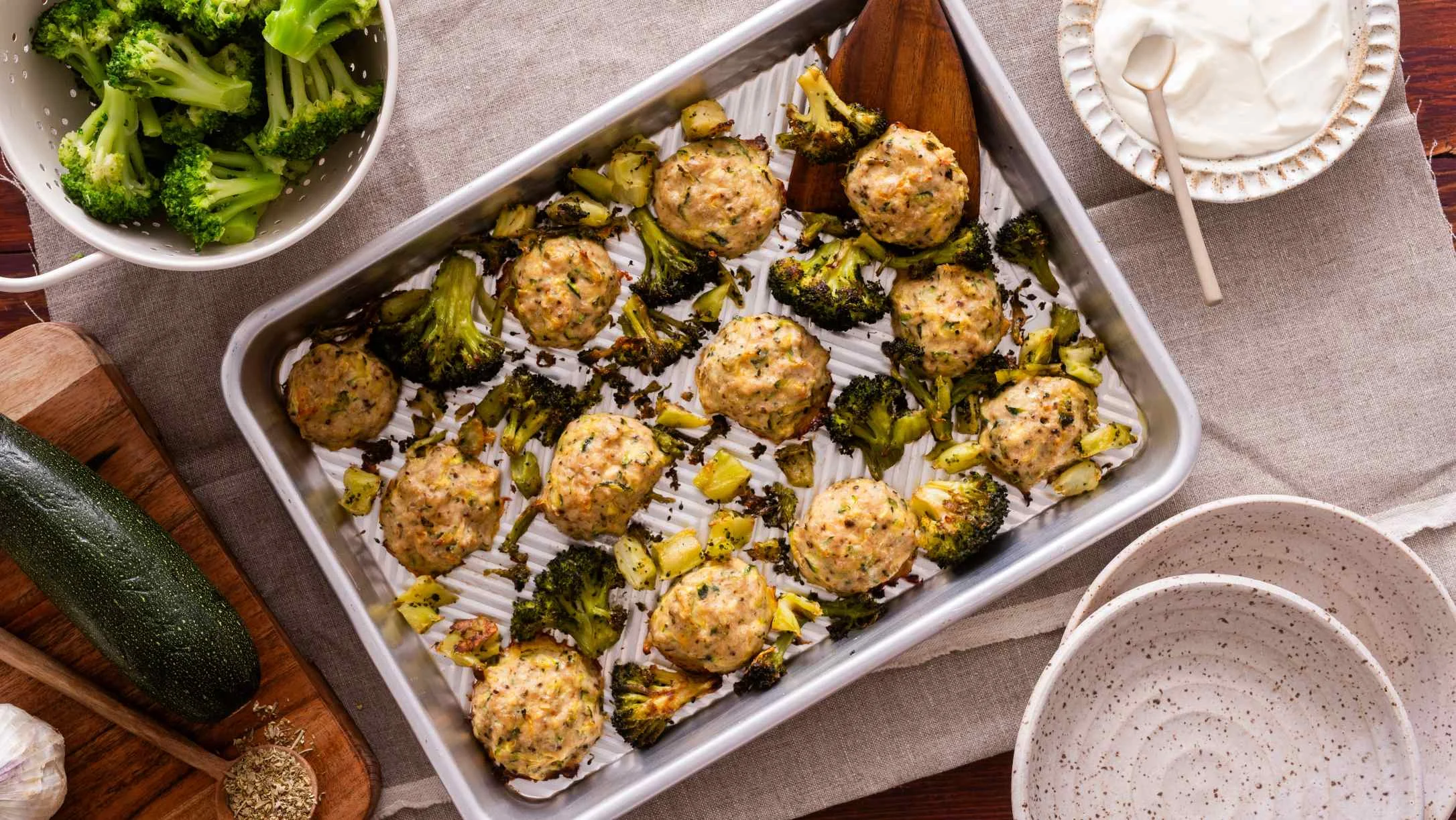 A baking pan with roasted chicken meatballs and broccoli, surrounded by fresh broccoli, zucchini, garlic, and a dipping sauce.