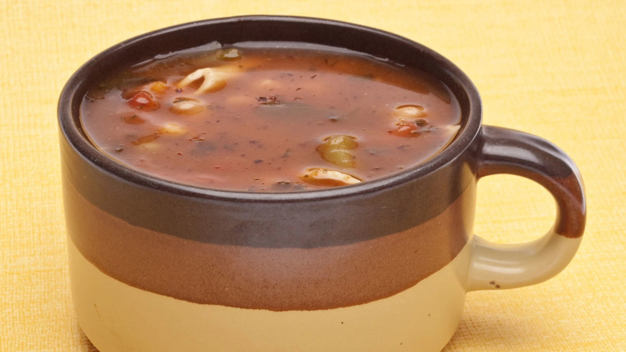 A brown ceramic mug filled with minestrone soup containing pasta and vegetables, set on a yellow textured surface.