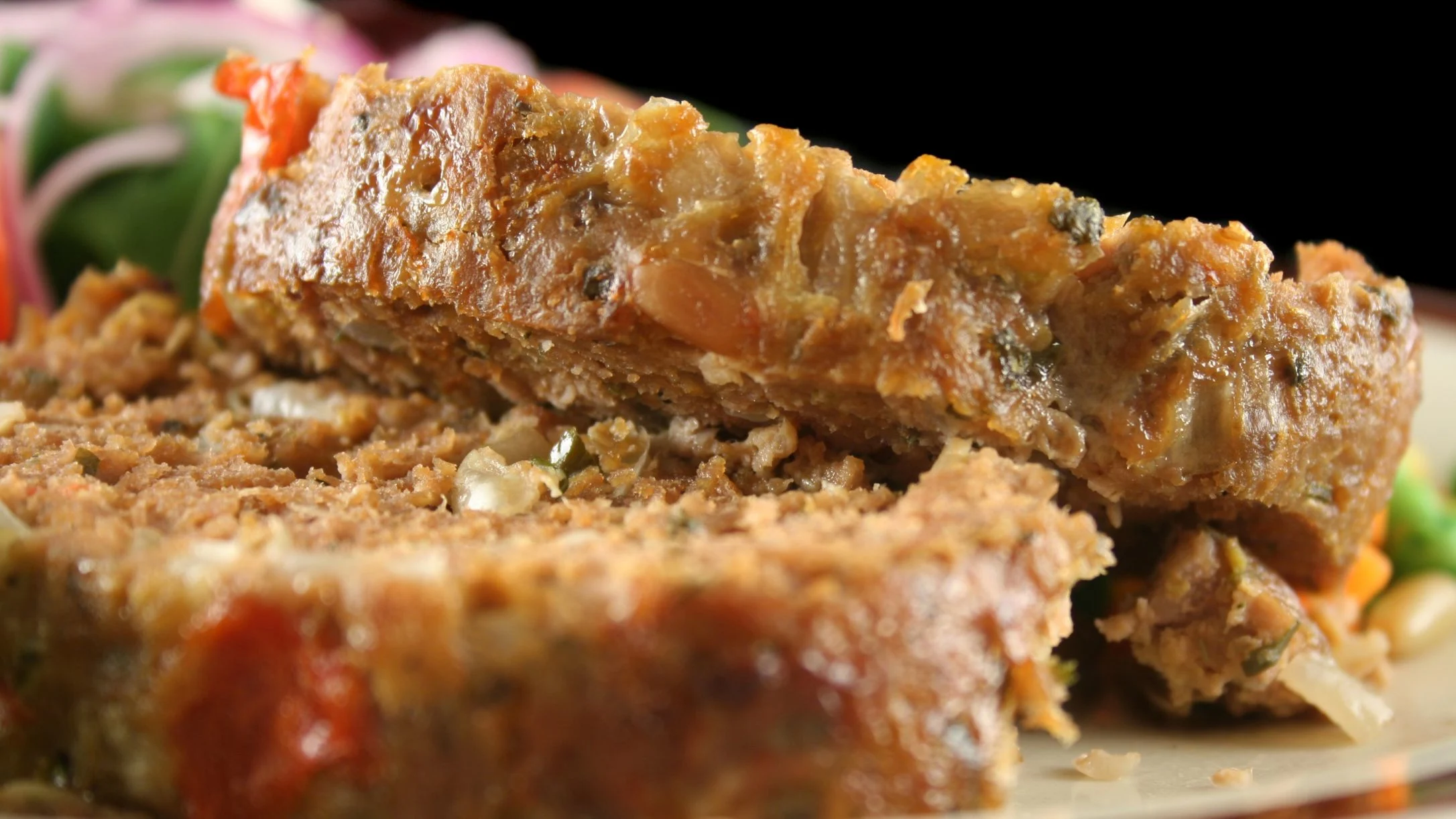 Close-up of two slices of meatloaf with vegetables, showing a golden-brown crust and moist interior, served on a plate.