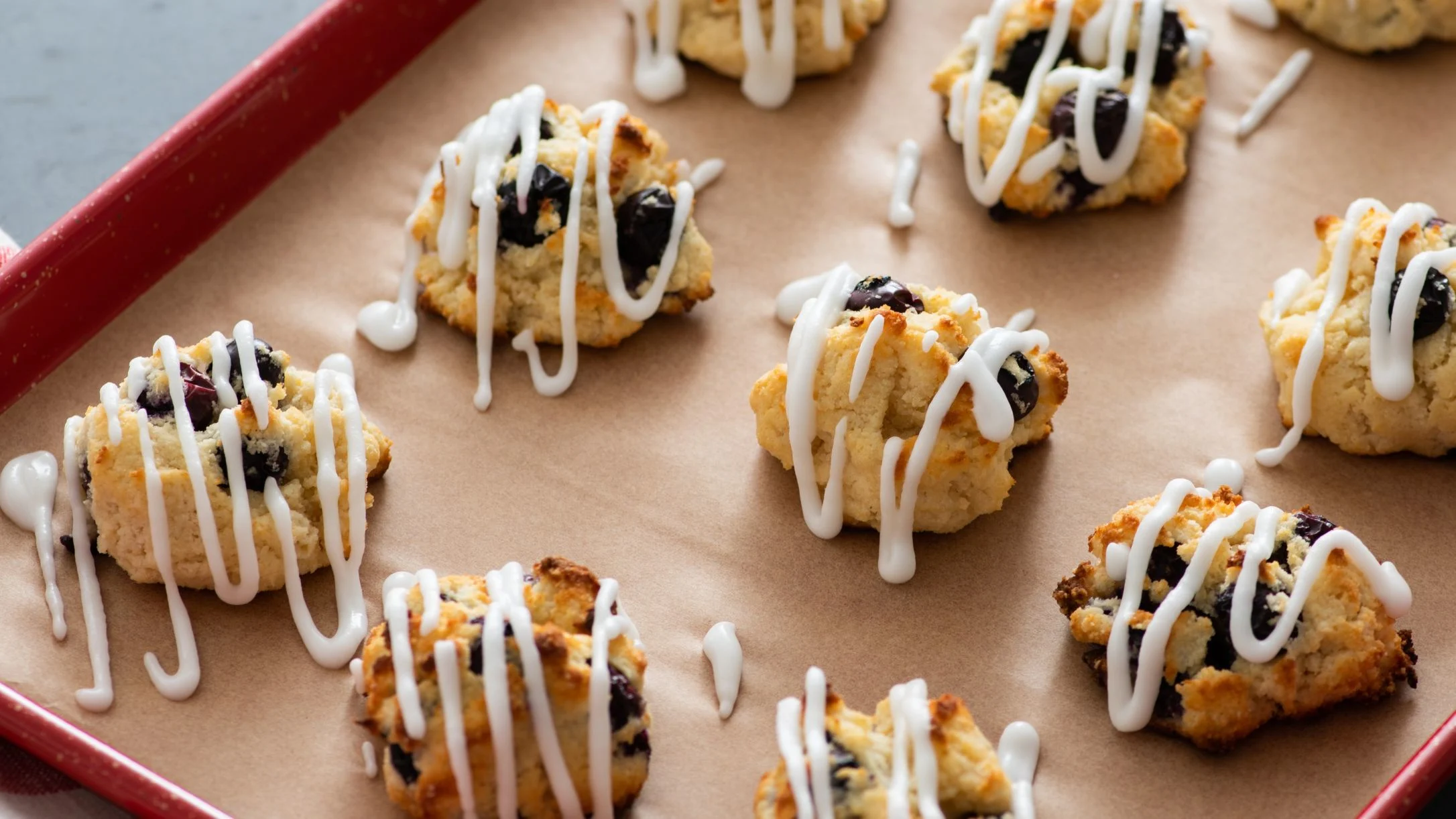 Freshly baked blueberry scones with white icing drizzle arranged on parchment paper in a red baking pan.