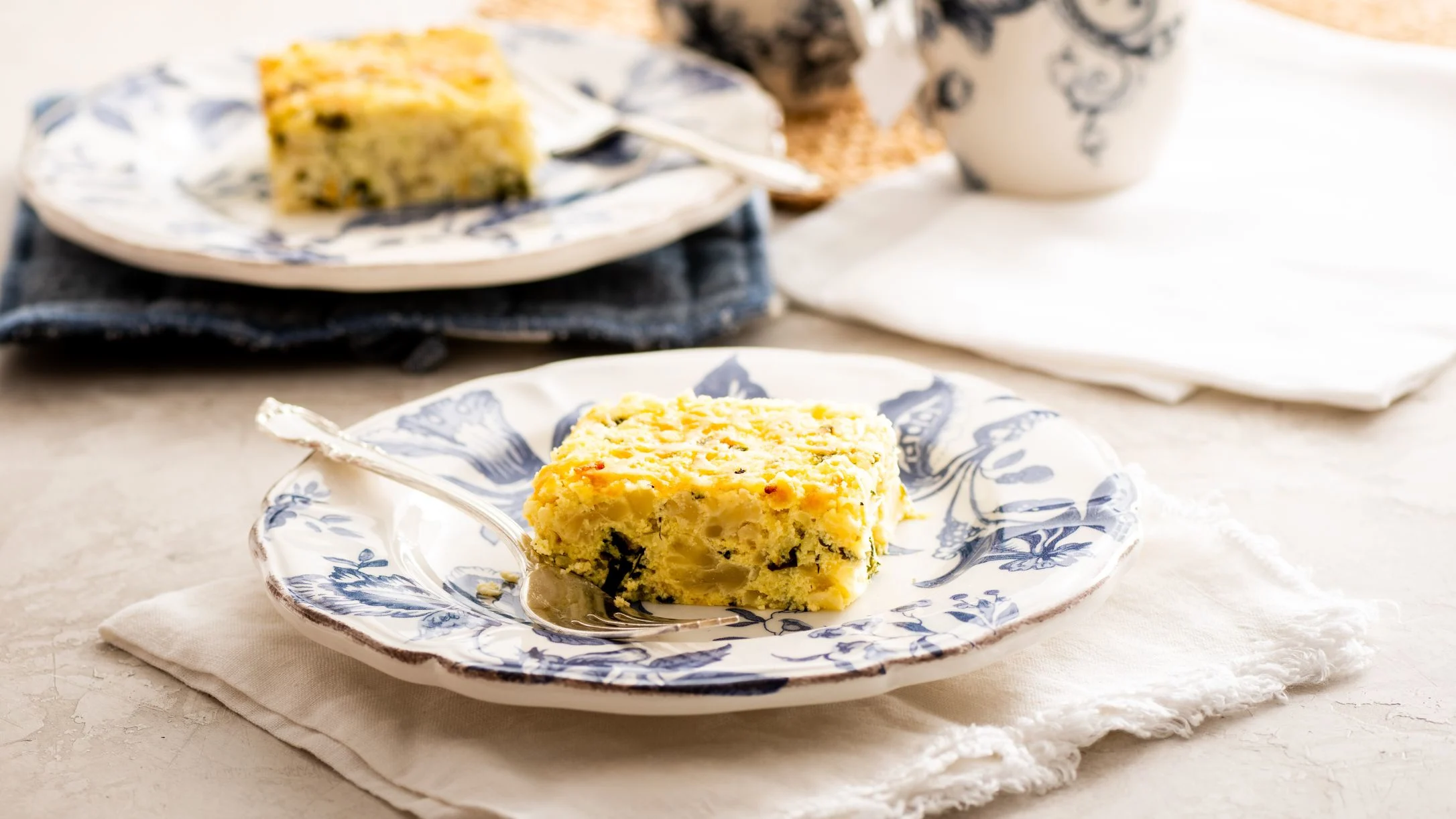 Slices of savory egg and cauliflower kugel with herbs served on blue and white decorative plates with forks on cream napkins.