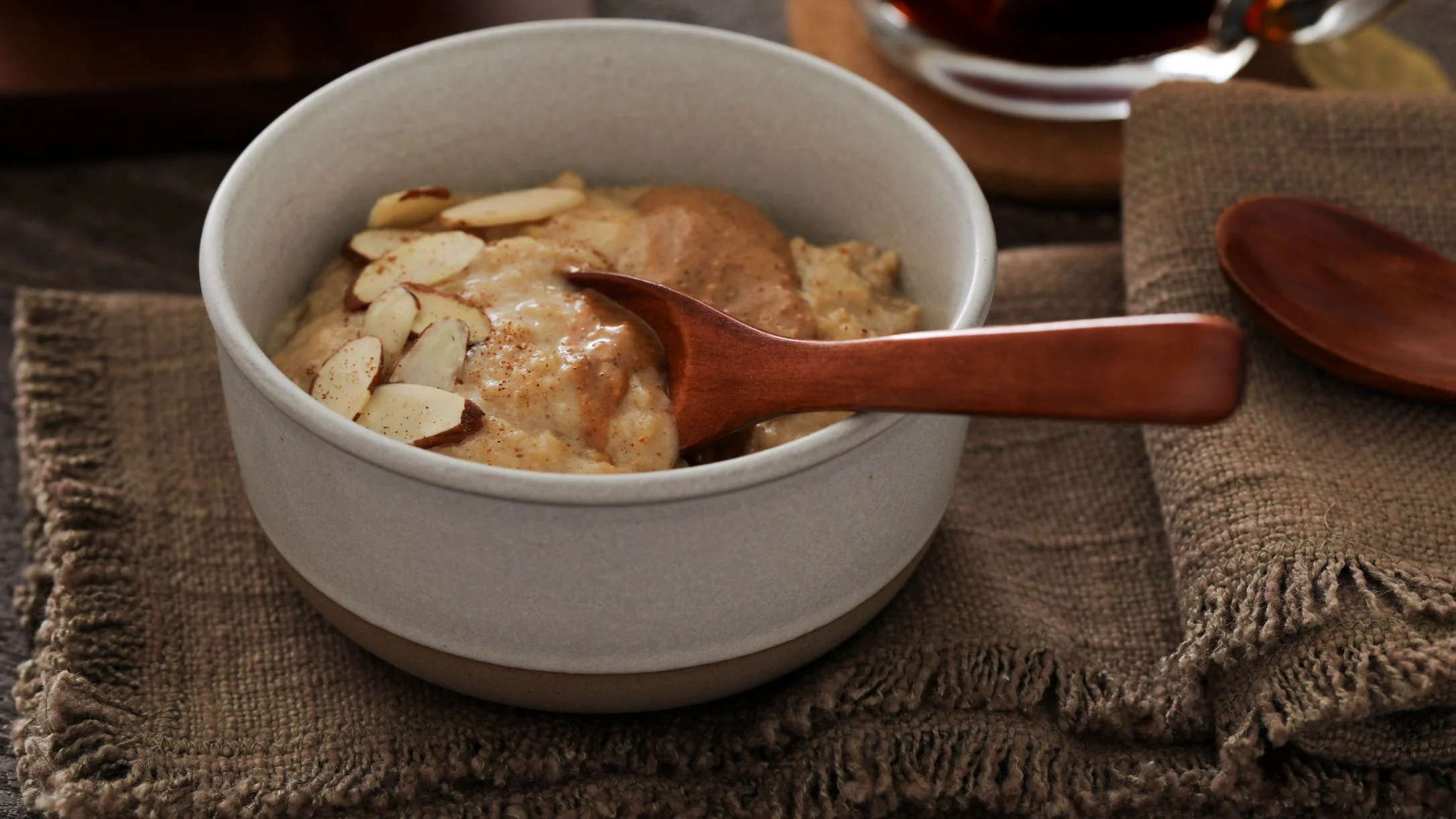 Bowl of oatmeal topped with sliced almonds and cinnamon, served with wooden spoon on textured brown cloth.