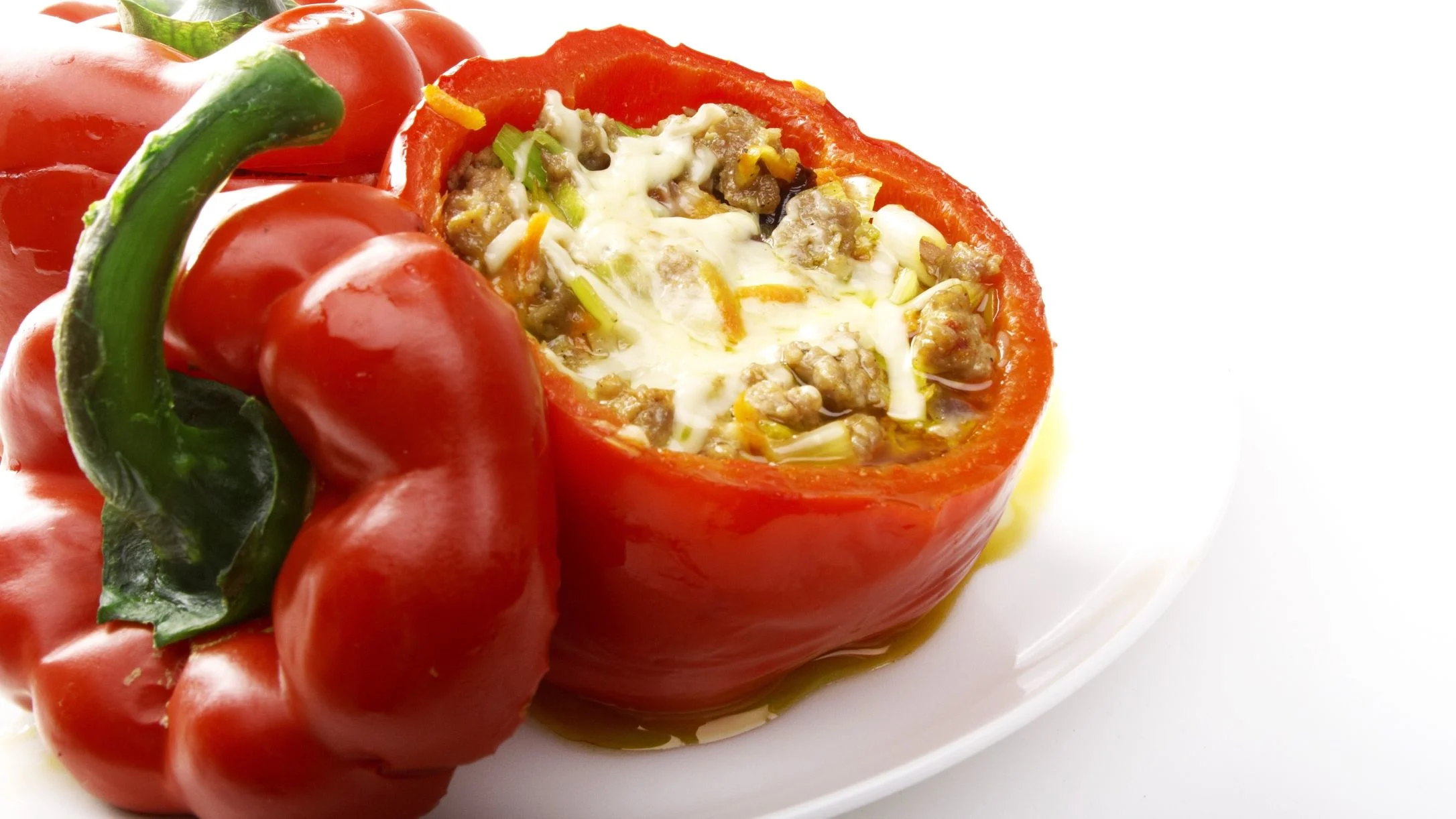 A red bell pepper stuffed with ground meat, vegetables, and melted cheese on a white plate, with whole red peppers nearby.