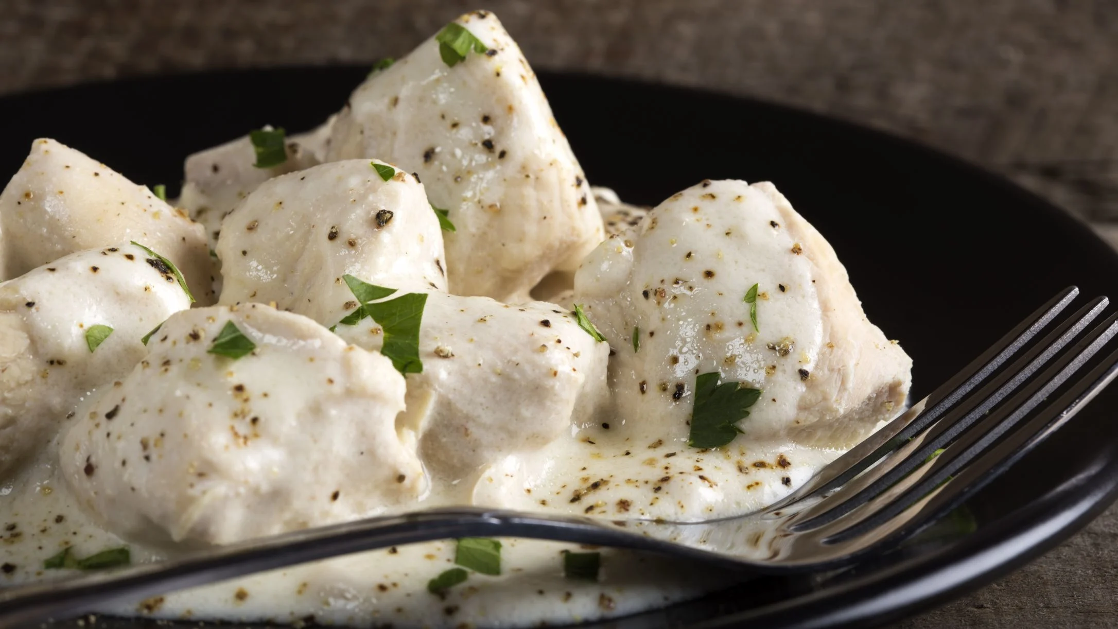 Chicken pieces in creamy white pepper sauce, garnished with fresh parsley, served on a black plate with a fork.