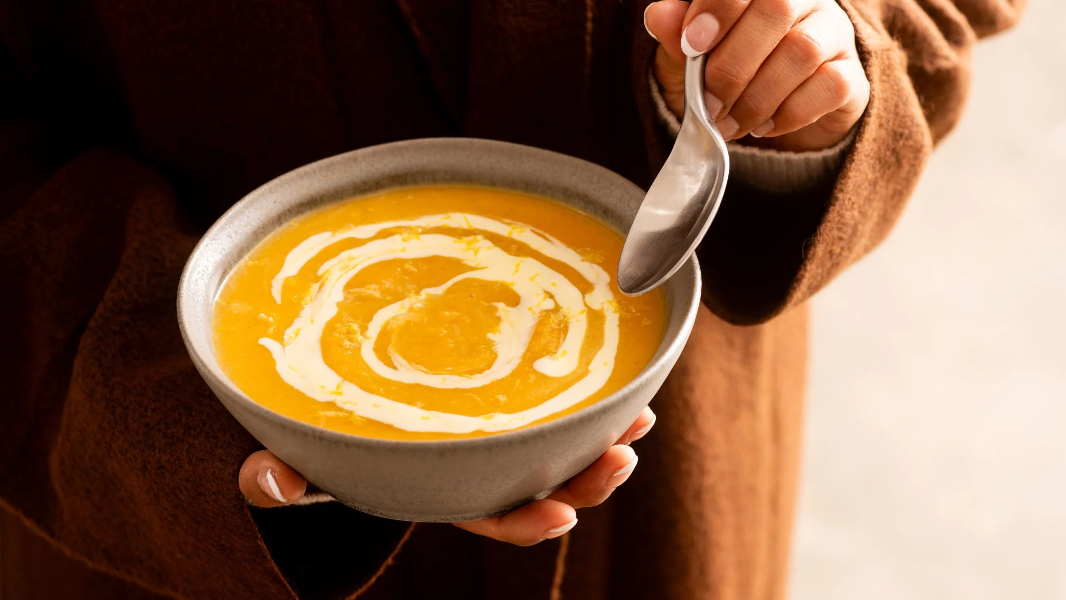 Hands holding a gray ceramic bowl of creamy orange butternut squash soup with a swirl of cream on top.