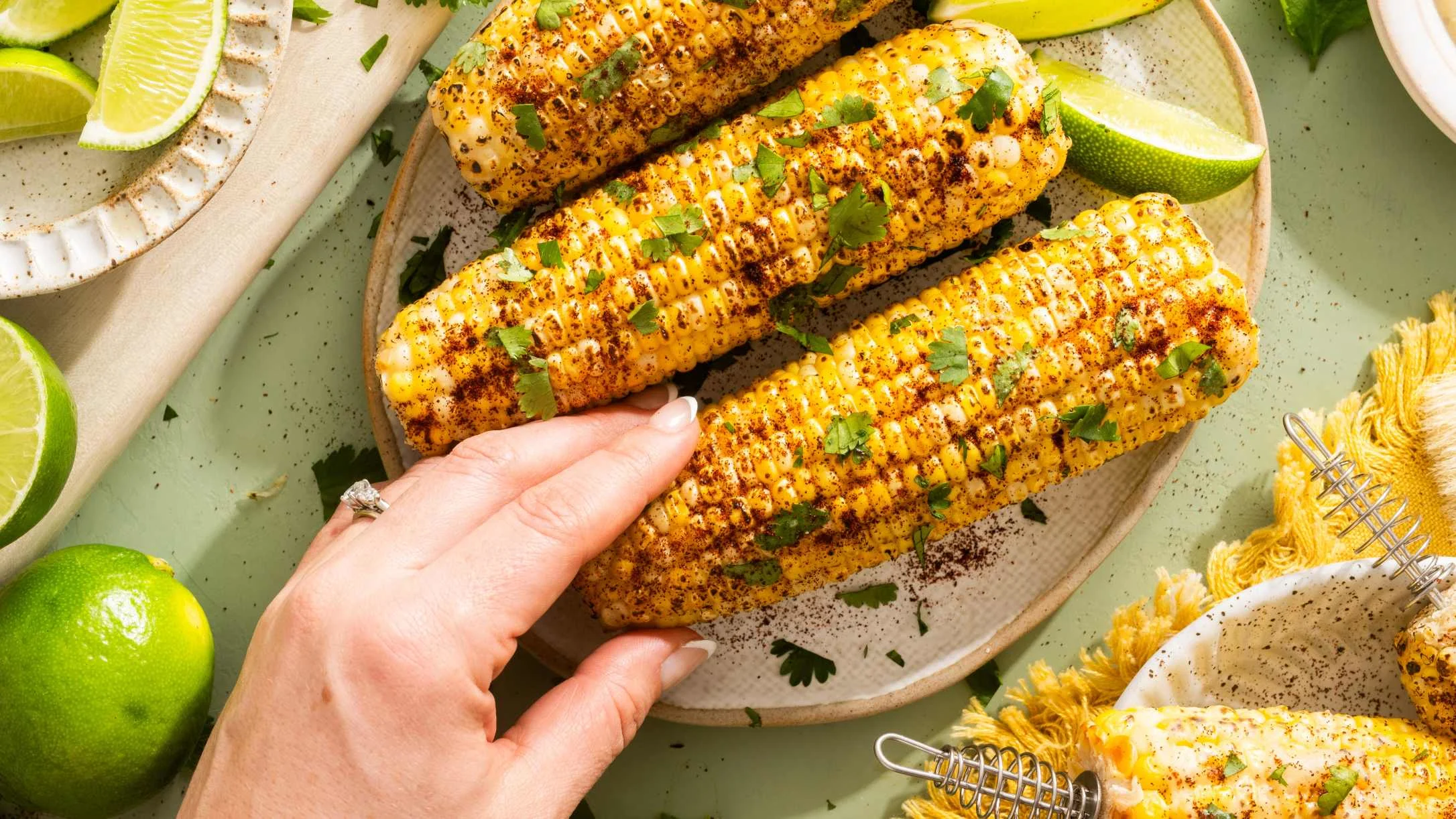 Grilled corn on the cob with spices and herbs on a ceramic plate, surrounded by lime wedges, with a hand reaching for one.