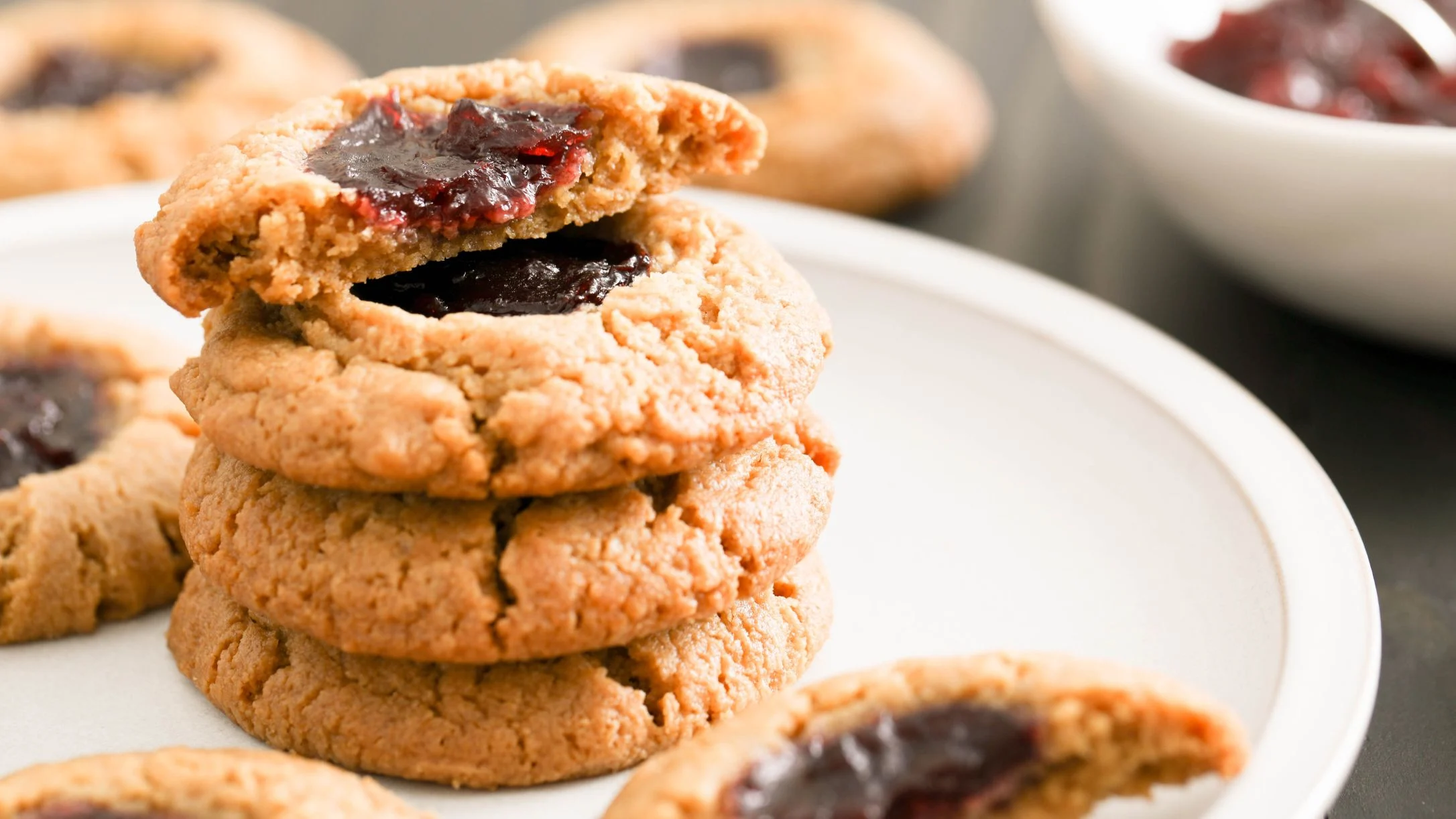 Stack of homemade thumbprint cookies with dark berry jam centers on a white plate, one cookie broken in half.