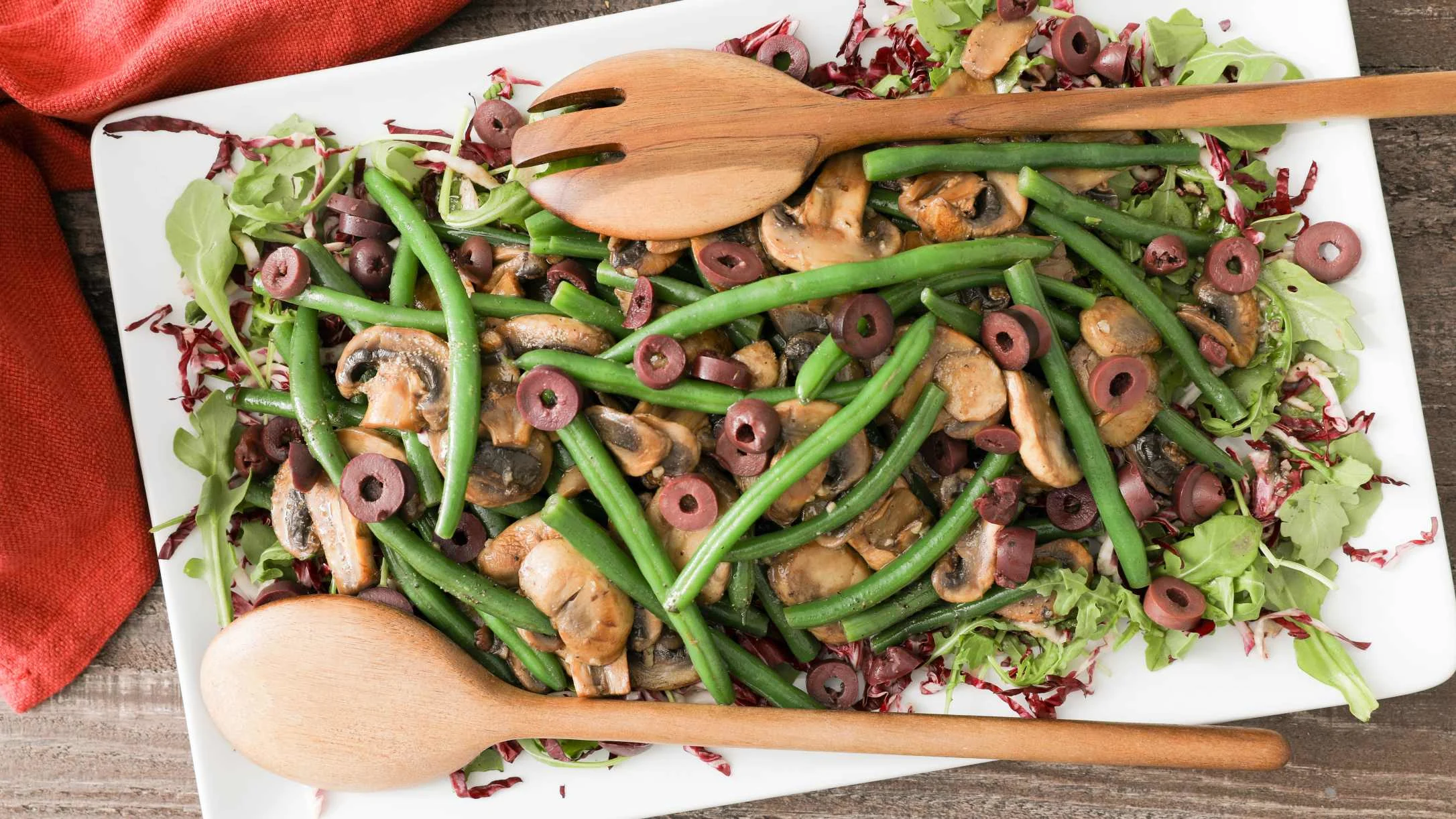 Fresh salad with green beans, mushrooms, and olives on a white platter with wooden serving utensils.