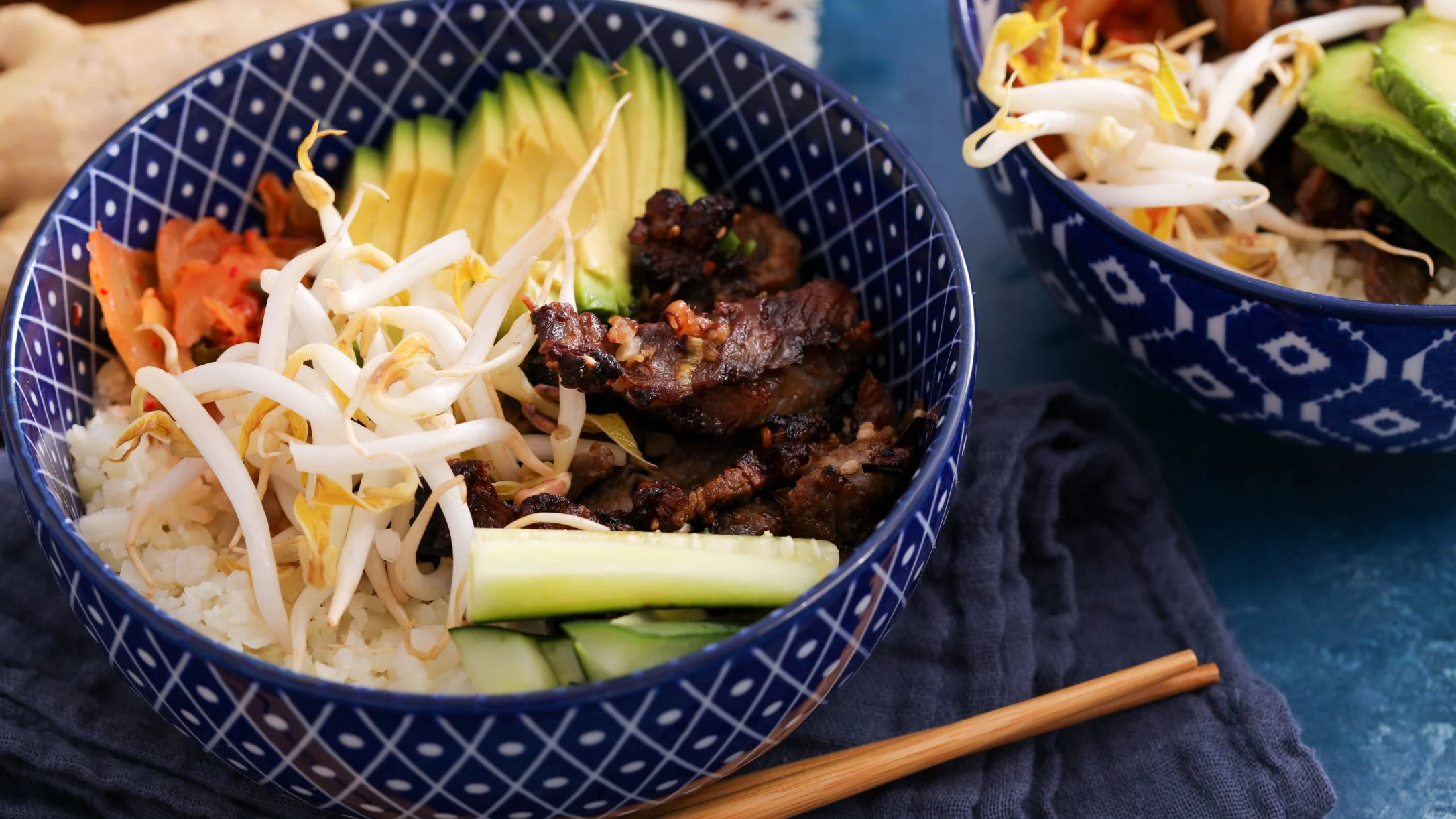 A blue patterned bowl with rice, grilled beef, avocado, bean sprouts, kimchi, and cucumber, served with chopsticks.