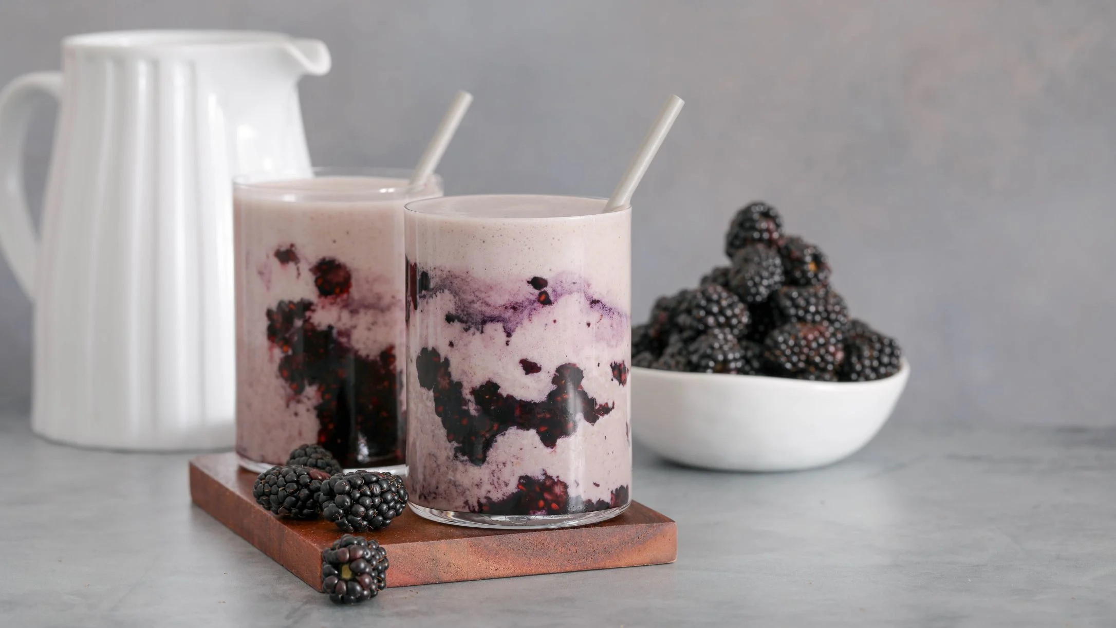 Two blackberry smoothies with straws on a wooden board, fresh blackberries, and a white pitcher on a gray surface.