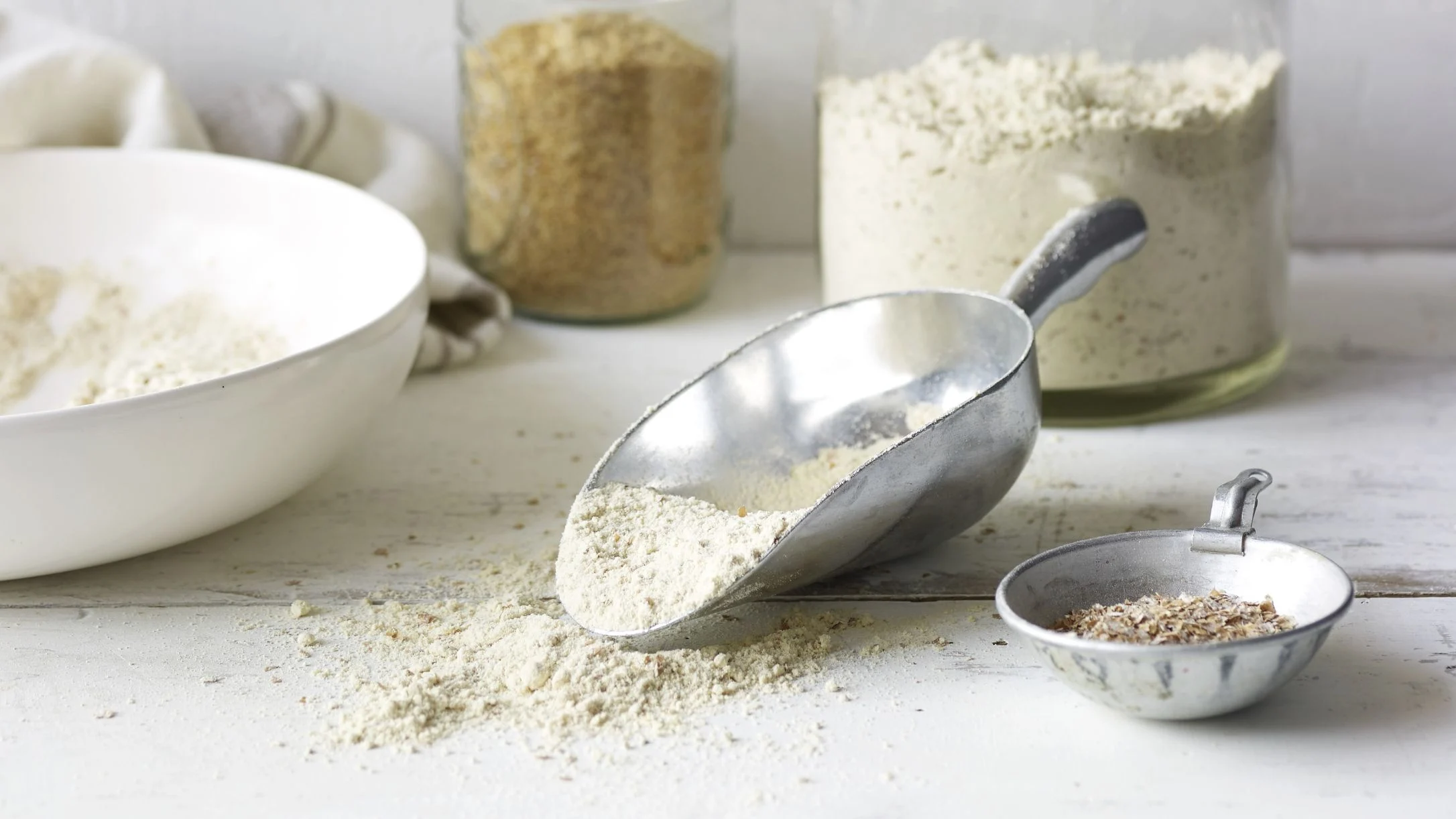 A metal scoop filled with flour on a white surface, with jars of flour, a white bowl, and a small measuring cup nearby.