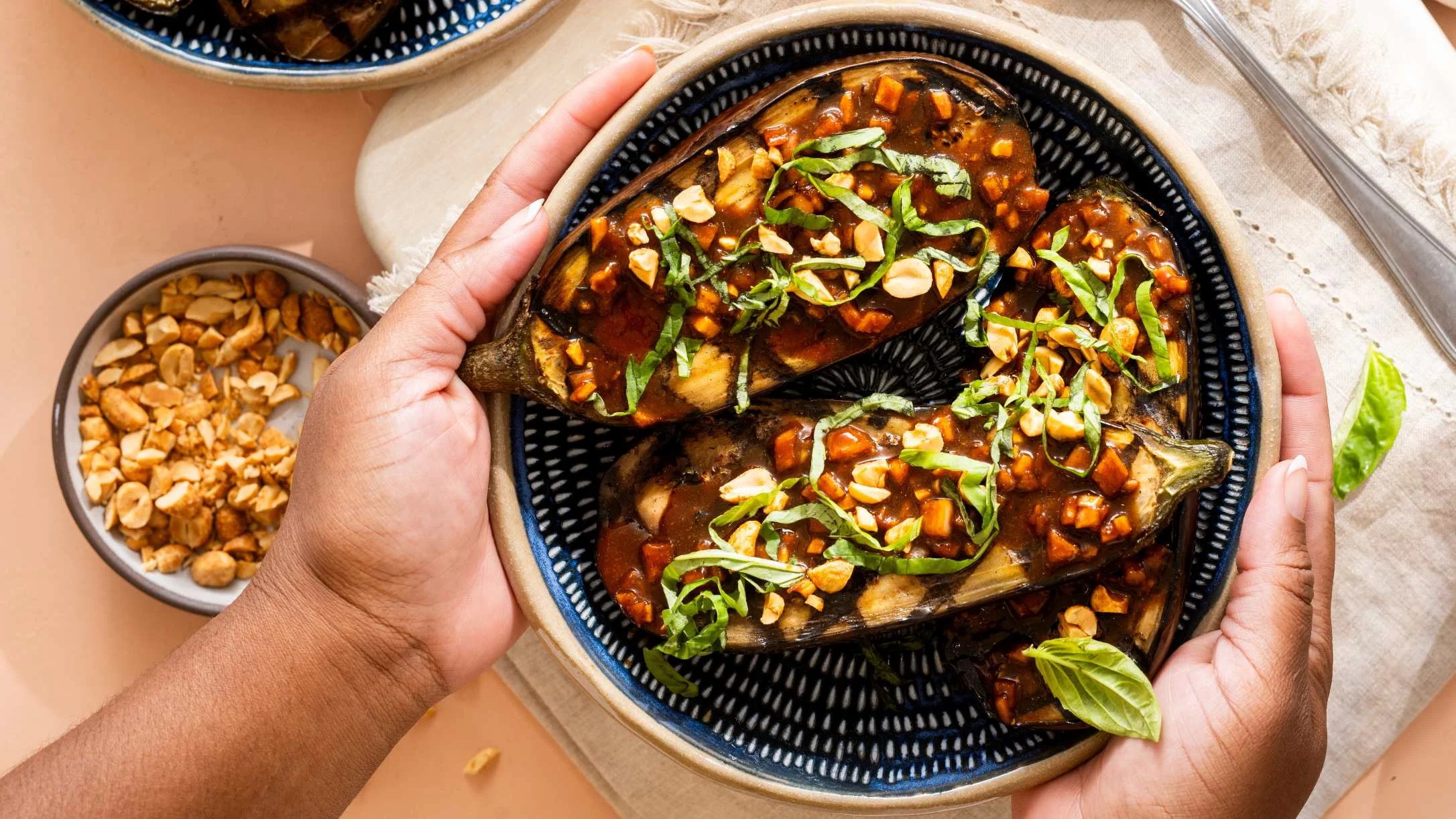 Hands holding a blue patterned plate with roasted eggplant halves topped with savory sauce, chopped peanuts, and fresh basil.