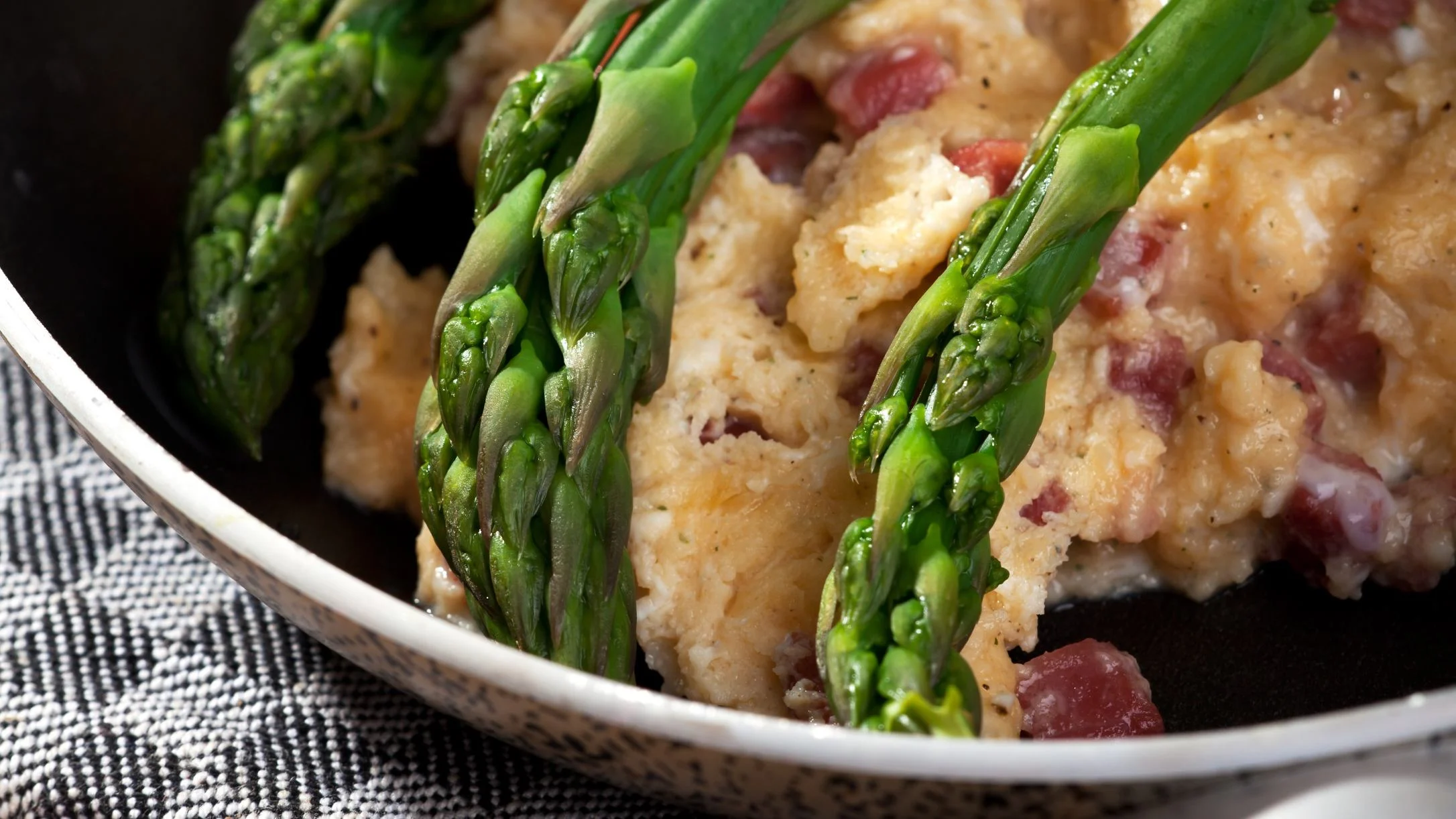 Close-up of a skillet dish featuring green asparagus spears over creamy scrambled eggs with diced bacon in a white pan.