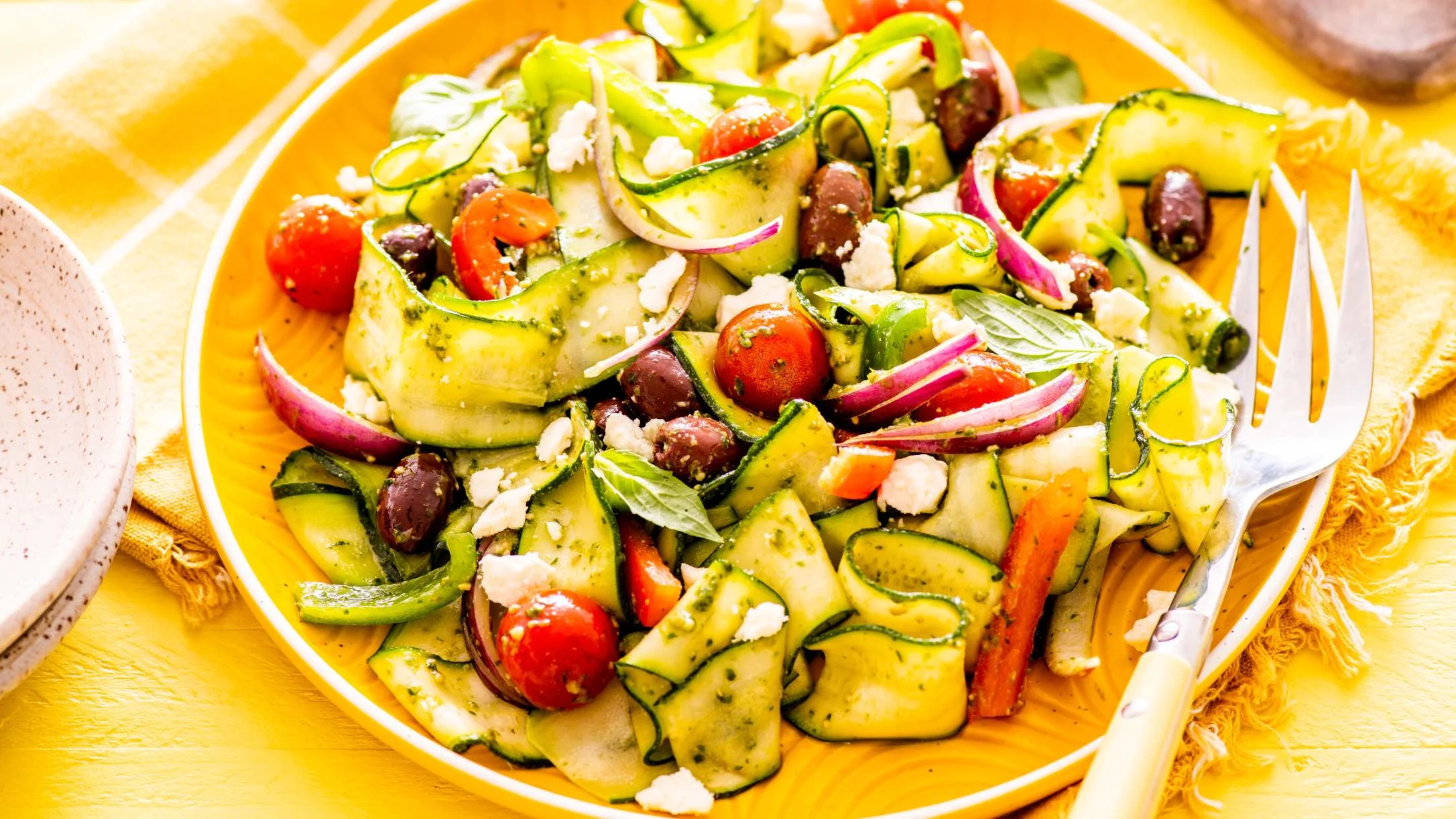 Fresh Mediterranean salad with zucchini ribbons, cherry tomatoes, olives, feta cheese, and red onions on a yellow plate.