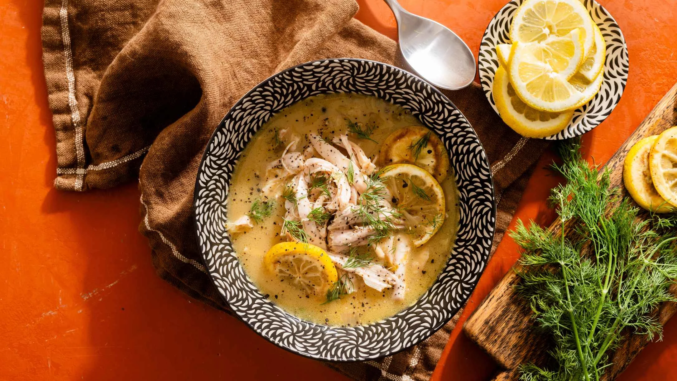 A bowl of creamy chicken soup with lemon slices and fresh dill, served in a patterned black and white bowl on an orange surface.