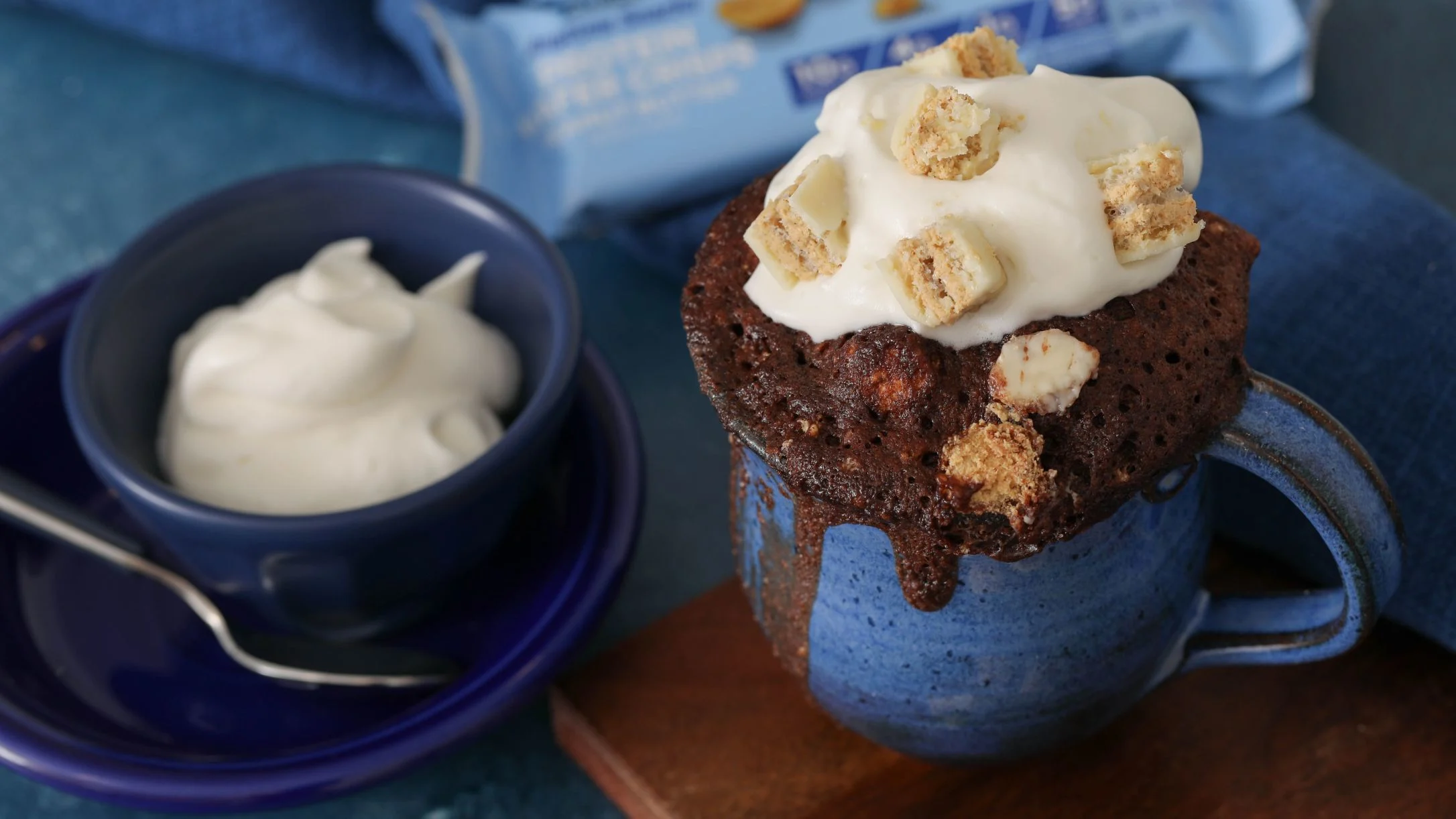 Chocolate mug cake topped with white cream and cookie crumbles, served alongside a blue bowl of whipped cream.