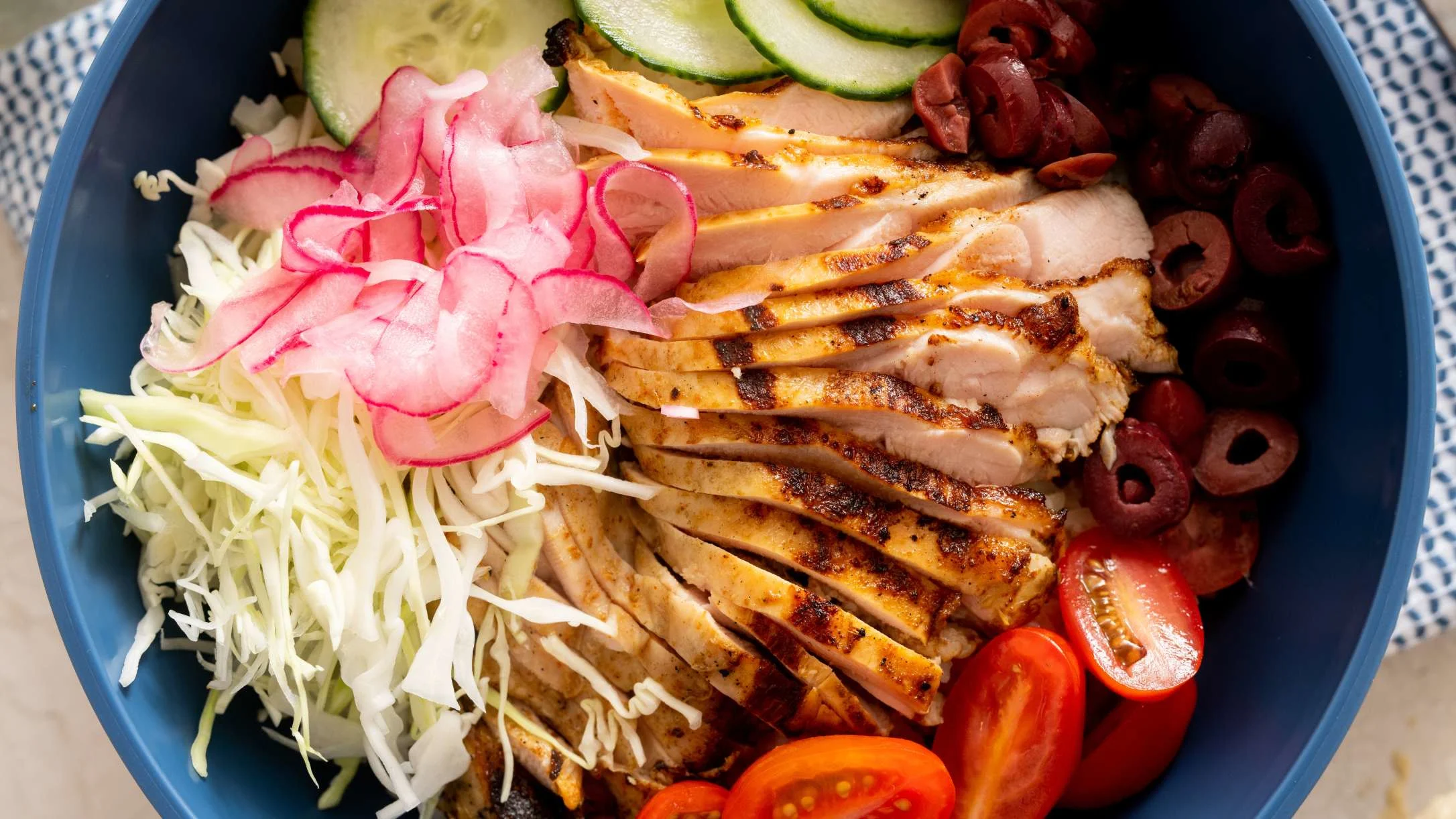 Blue bowl with sliced grilled chicken, shredded cabbage, pickled radish, cucumber, olives, and cherry tomatoes.