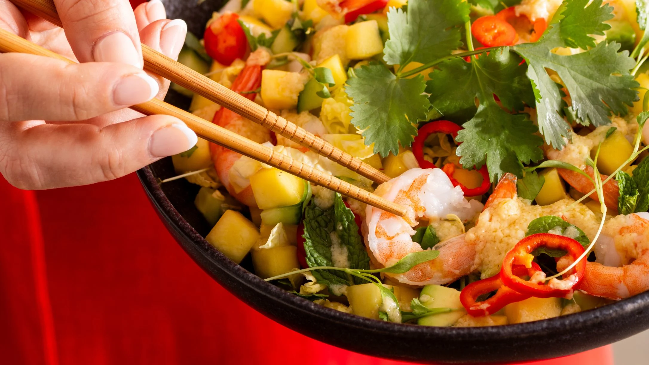 Hand using chopsticks to pick up shrimp from a colorful salad with vegetables, cilantro, and red chili slices.