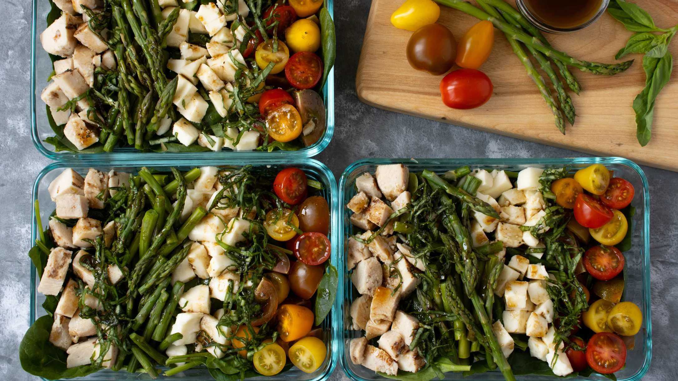 Three glass meal prep containers filled with grilled chicken, asparagus, mozzarella, and colorful cherry tomatoes.