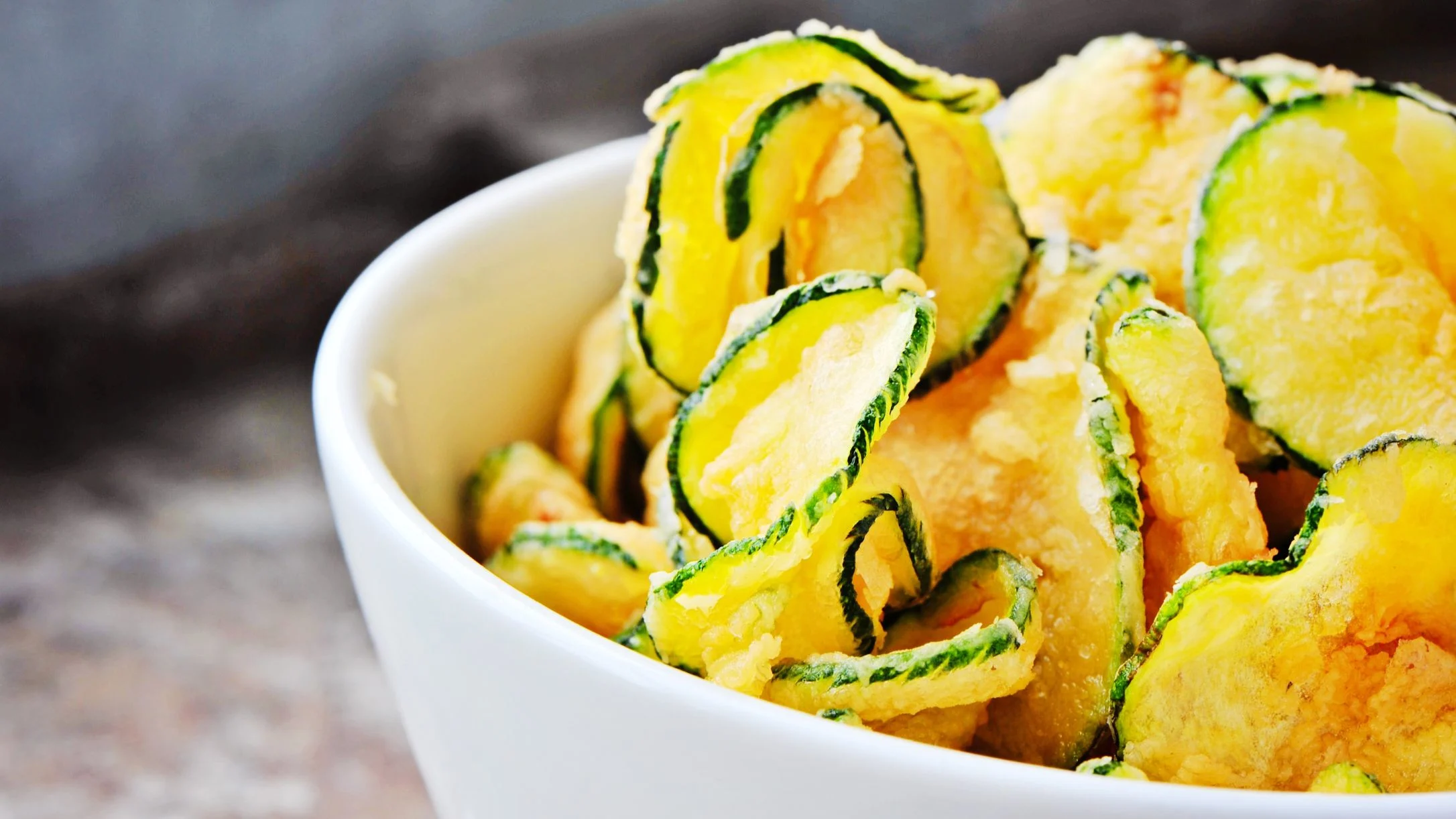 A white bowl filled with crispy zucchini chips, showing golden yellow and green edges, on a stone surface.