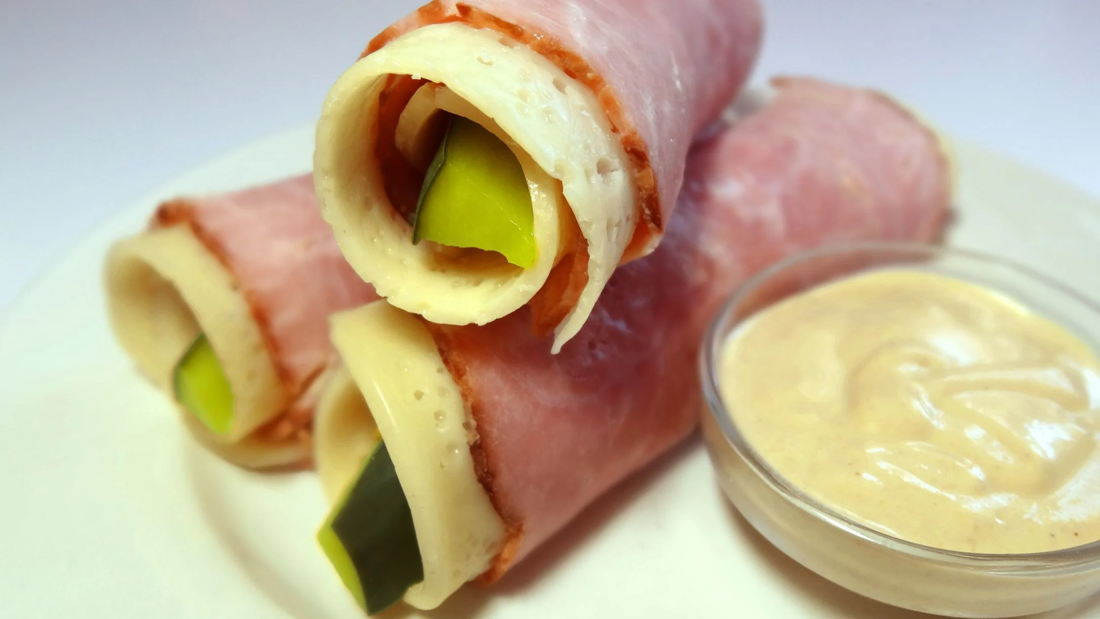 Ham and cheese rolls filled with pickles on a white plate, served with a creamy dipping sauce in a small glass bowl.