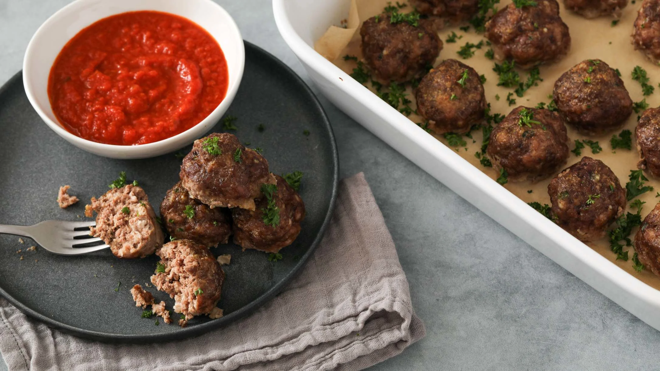 Homemade meatballs on a plate and in a baking dish, garnished with parsley, served with red marinara sauce.